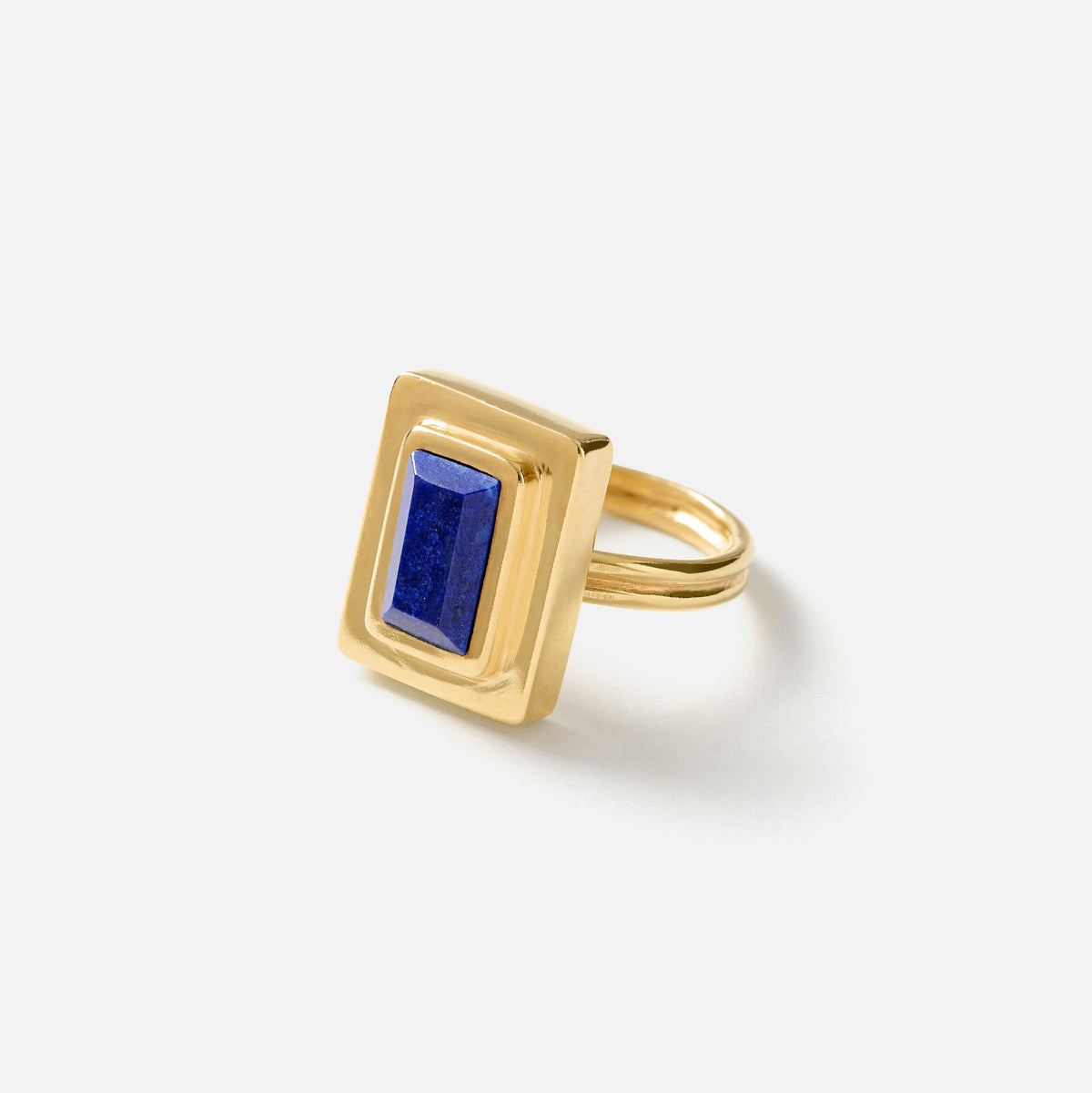 A gold ring featuring a rectangular bezel-set lapis lazuli stone, photographed on a clean, light gray background.