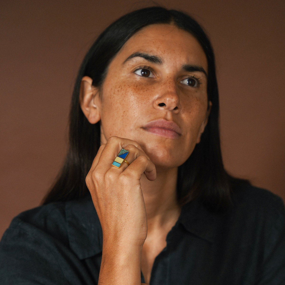 A close-up portrait of a woman with dark hair and freckles, wearing a dark shirt and resting her chin on her hand, which is adorned with a colorful geometric ring. The background is a solid warm brown.