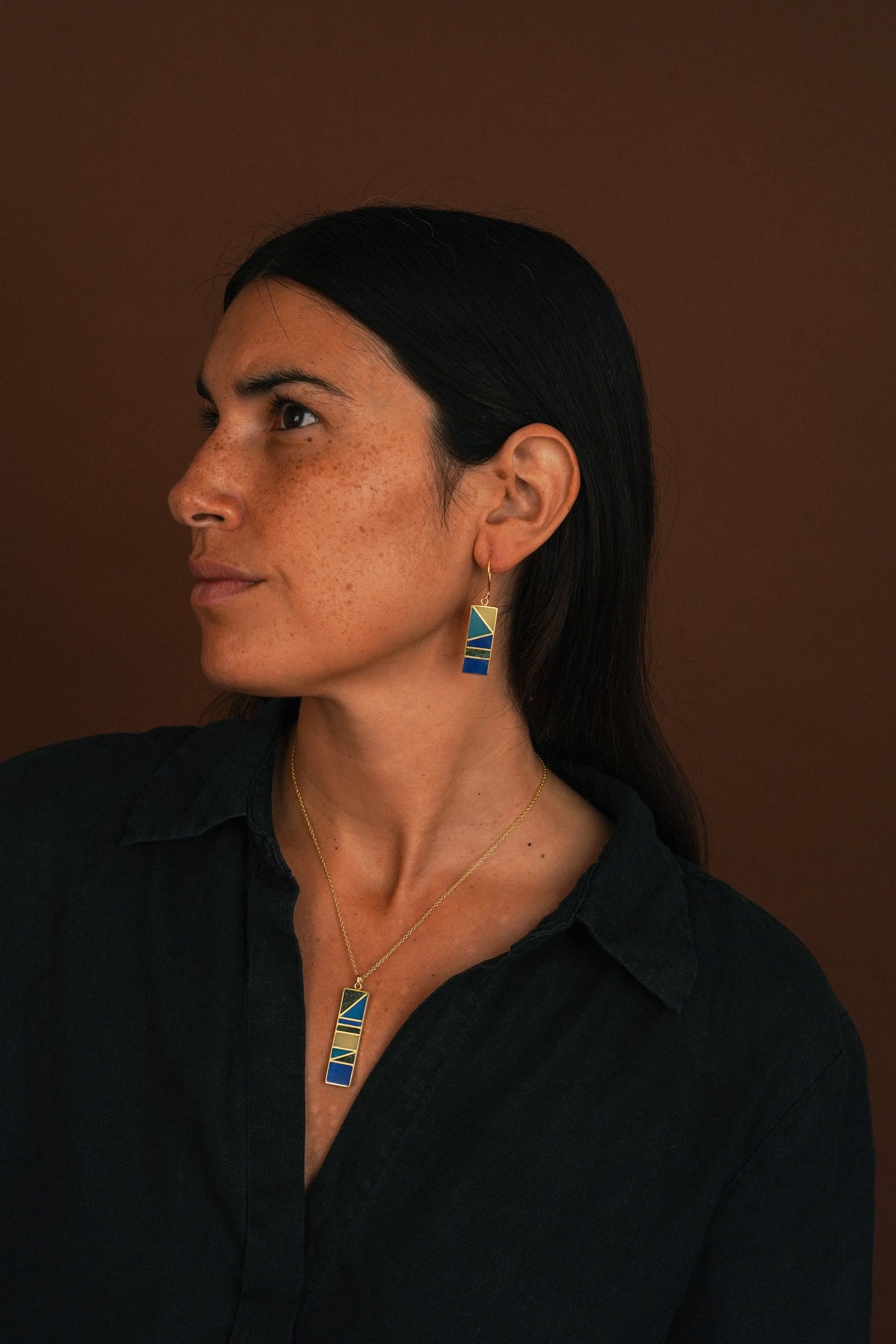 A woman in profile wearing a gold necklace and matching drop earrings with blue and yellow geometric enamel details, set against a solid dark brown background.
