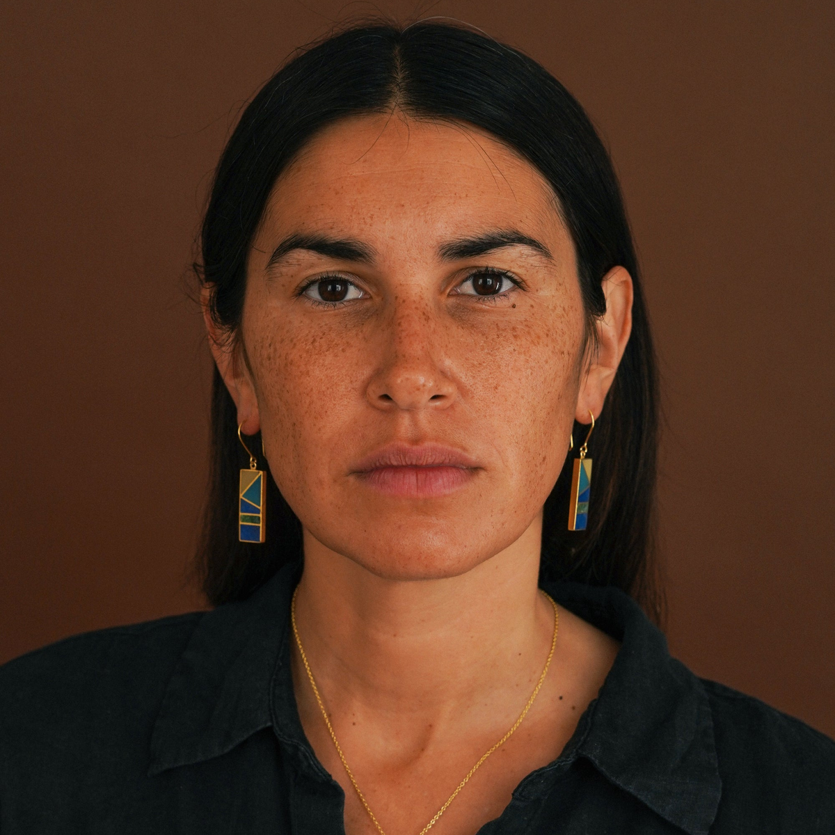 A close-up portrait of a woman with dark hair and freckles, wearing rectangular blue and gold geometric drop earrings and a gold necklace, against a solid brown background.
