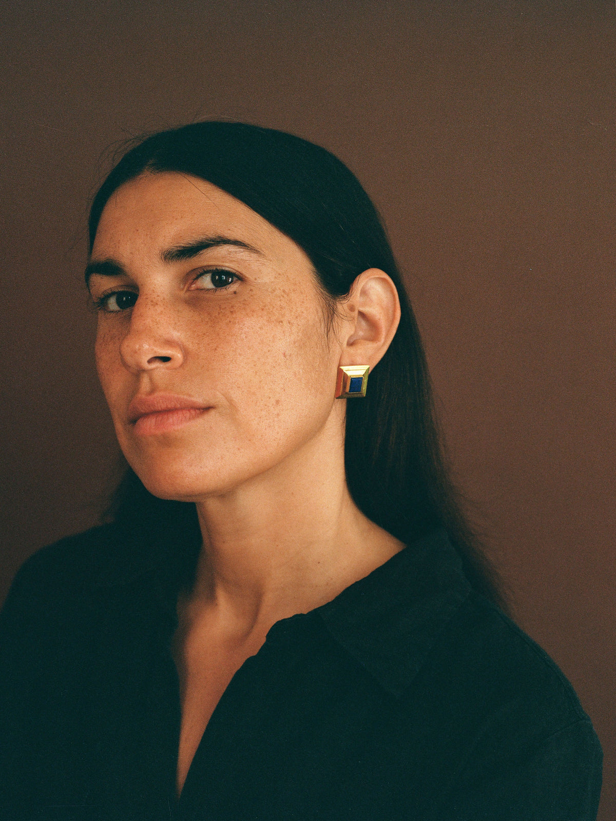 woman with dark hair wearing a dark shirt wears large gold earrings with lapis lazuli on a brown background