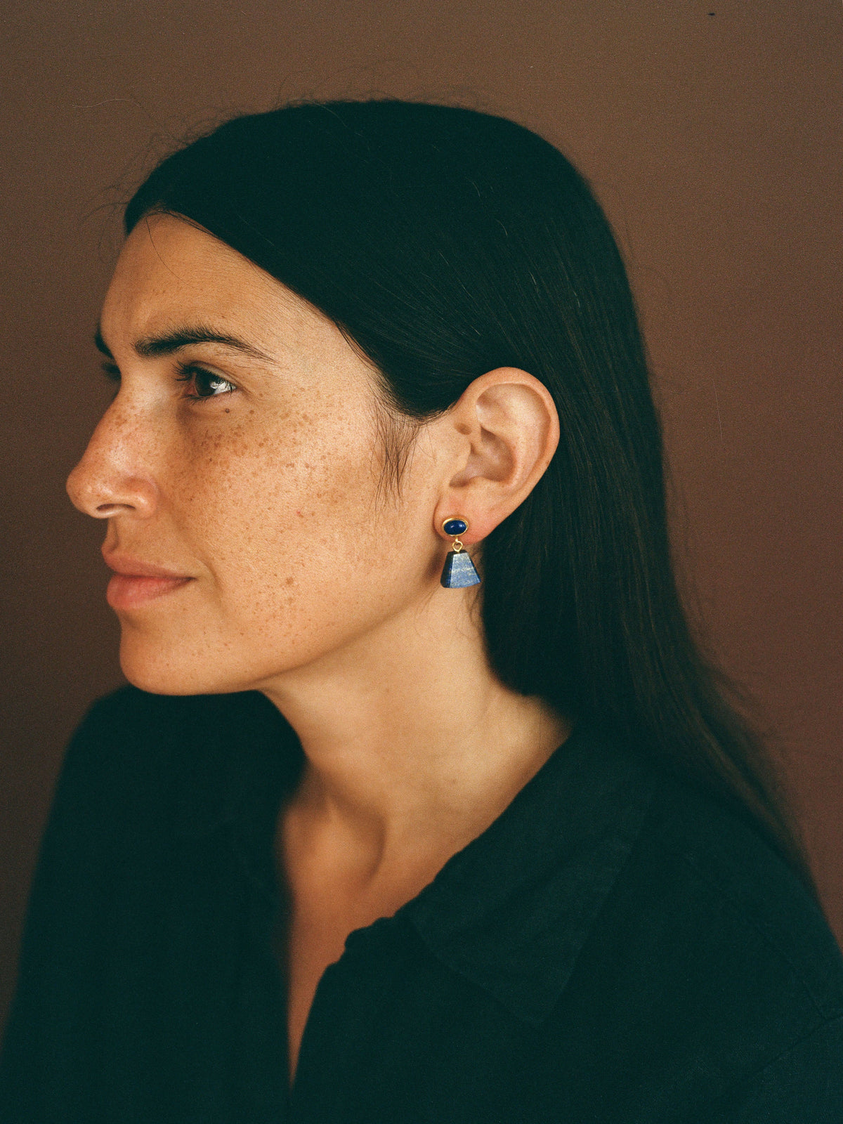 woman with dark hair in side profile wears dangling lapis lazuli earrings and a dark shirt on a brown background