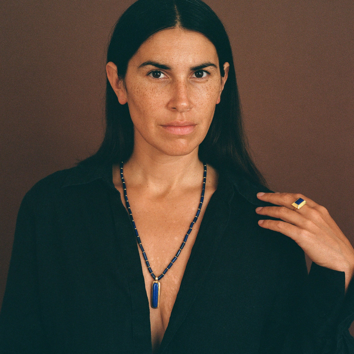 A woman with dark hair and freckles wears a long beaded necklace with a blue rectangular pendant and a gold ring on one finger, posed against a solid brown background.