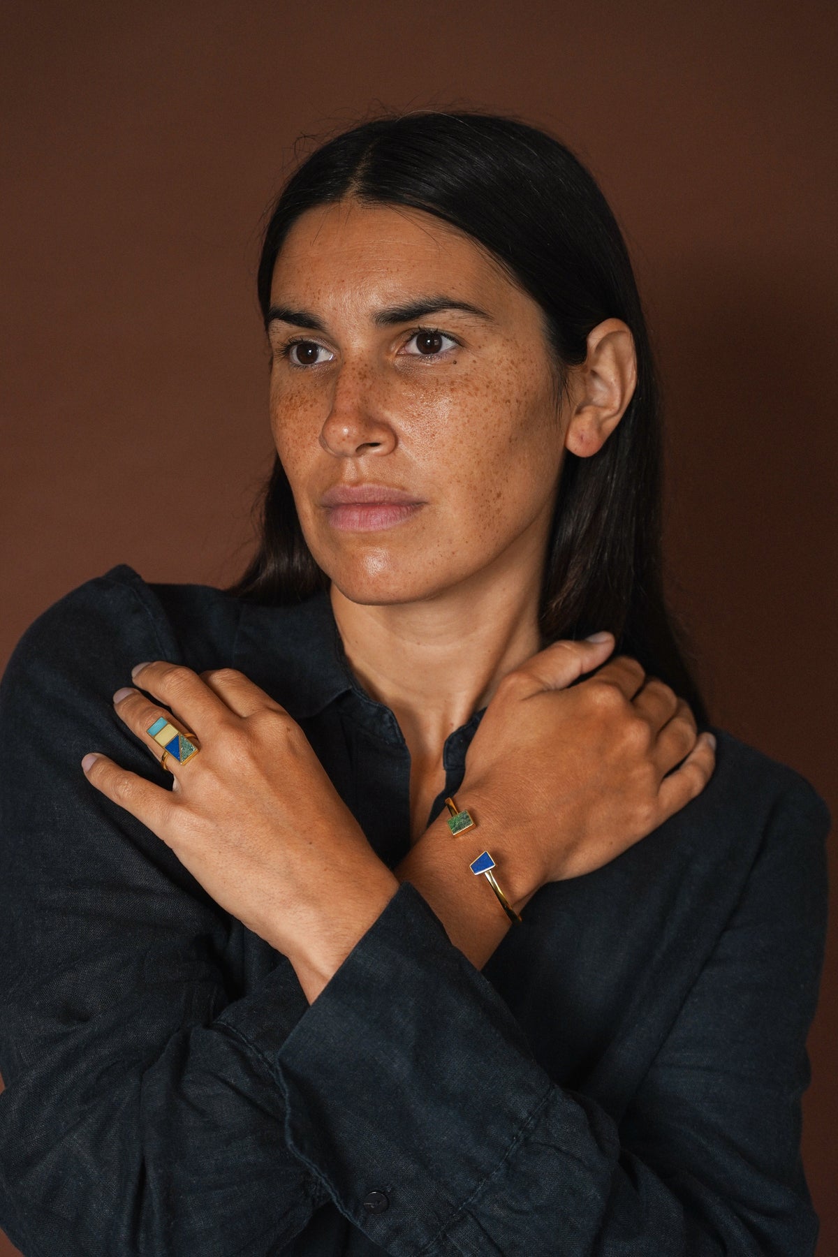 woman with crossed arms across her chest wearing a gold bangle and ring with semi-precious stones, on a brown background.