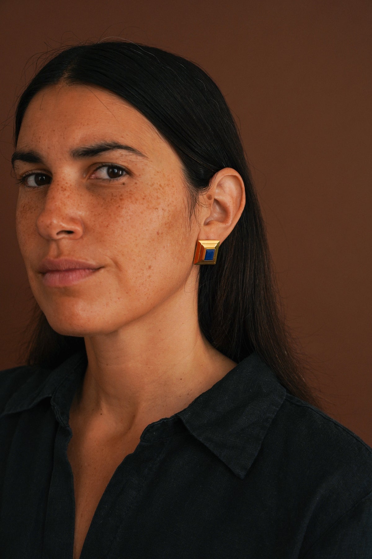 A close-up portrait of a woman with dark hair wearing a small geometric gold and blue stud earring, set against a solid warm brown background.