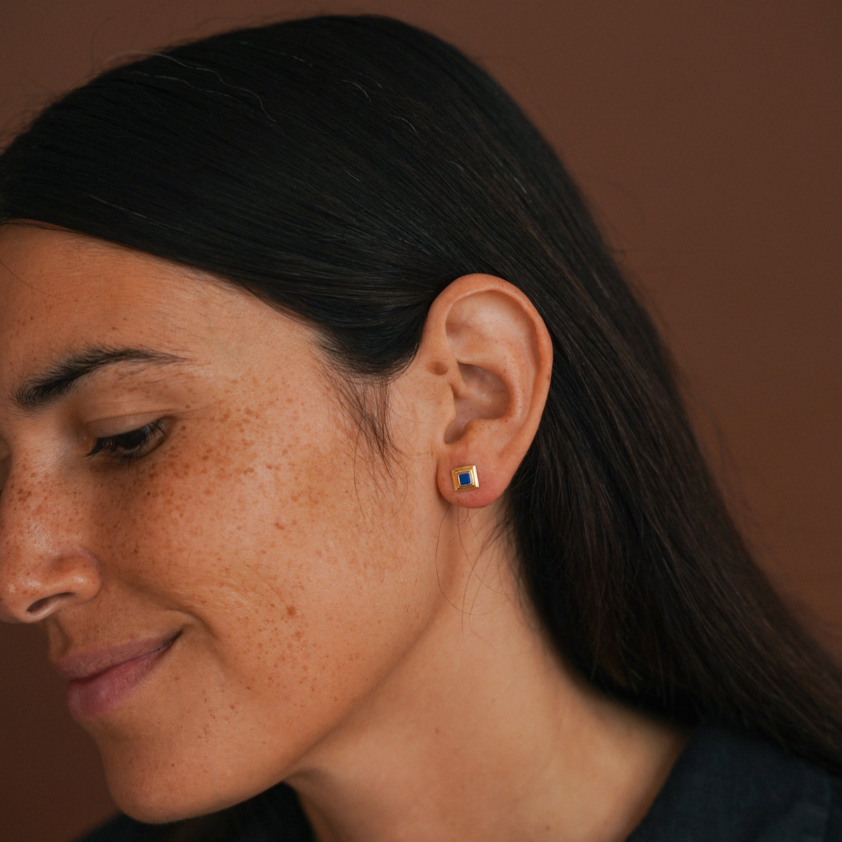 A close-up of a woman's ear wearing a small square stud earring with a colorful flag design, set against a warm brown background.
