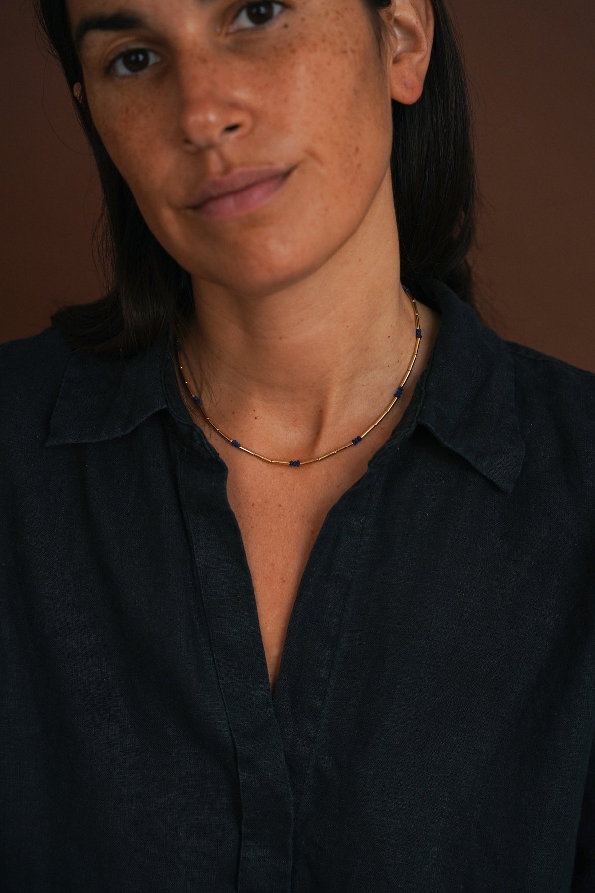 A close-up of a woman wearing a delicate gold chain necklace with small dark blue bead accents, paired with a dark navy button-down shirt against a warm brown background.