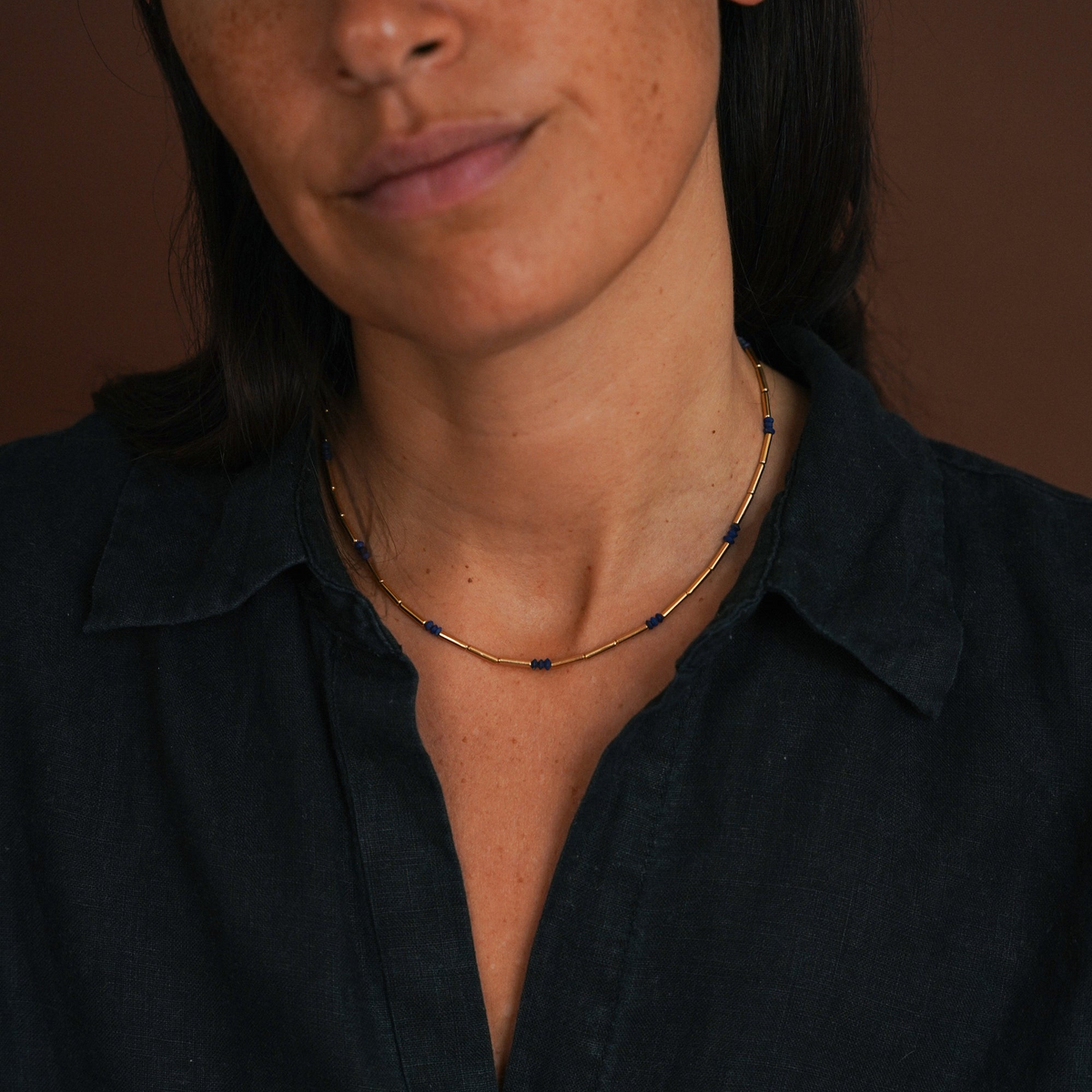 A close-up of a person wearing a delicate gold chain necklace with small dark blue bead accents, paired with a dark navy shirt against a warm brown background.