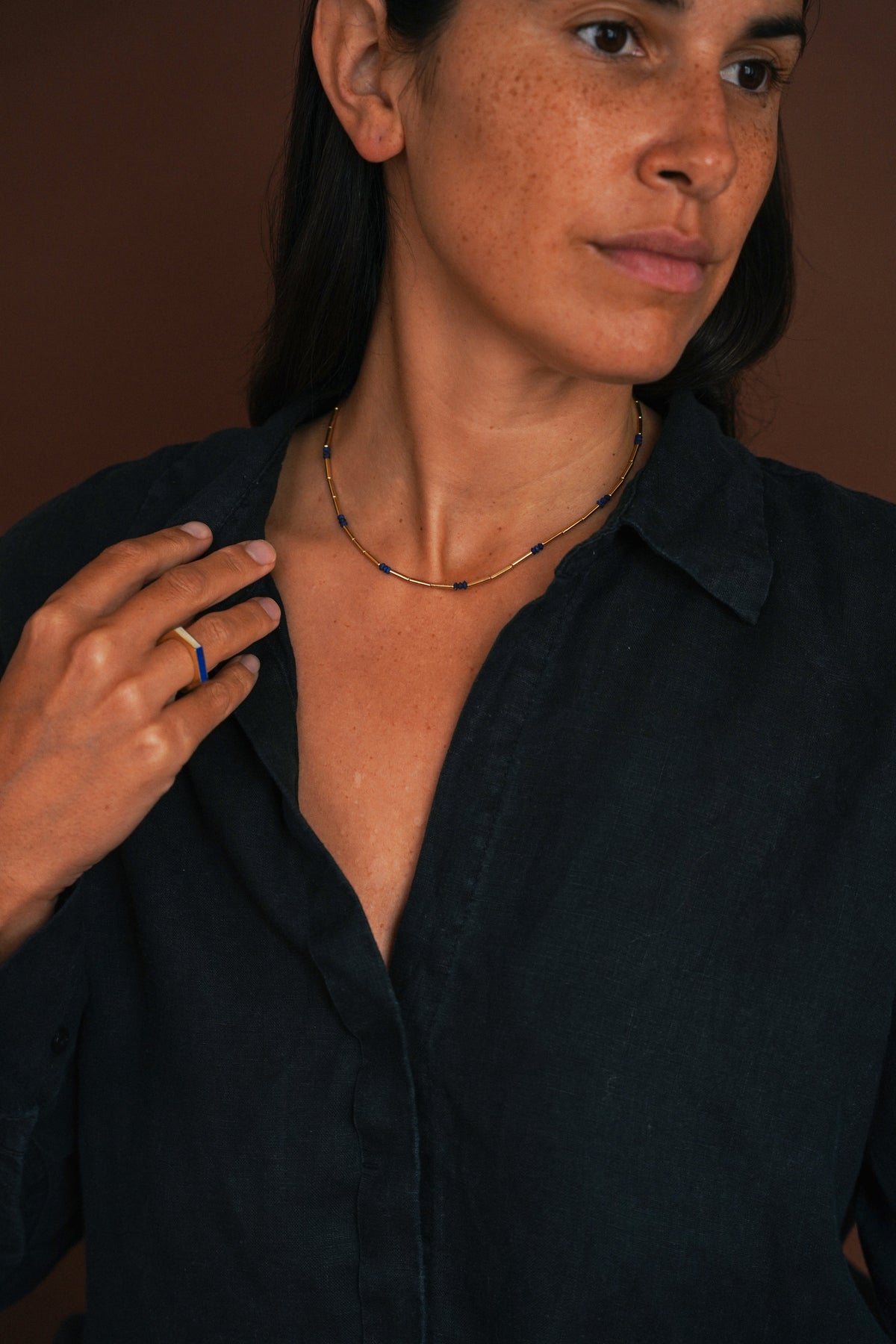 woman with dark hair wearing ring with stones and a beaded chain necklace and a dark shirt on a brown background