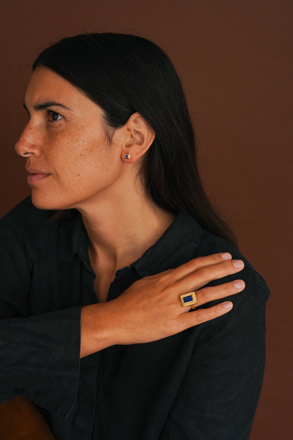 A woman in a dark shirt wearing a gold ring with a blue rectangular stone, shown in profile against a warm brown background.