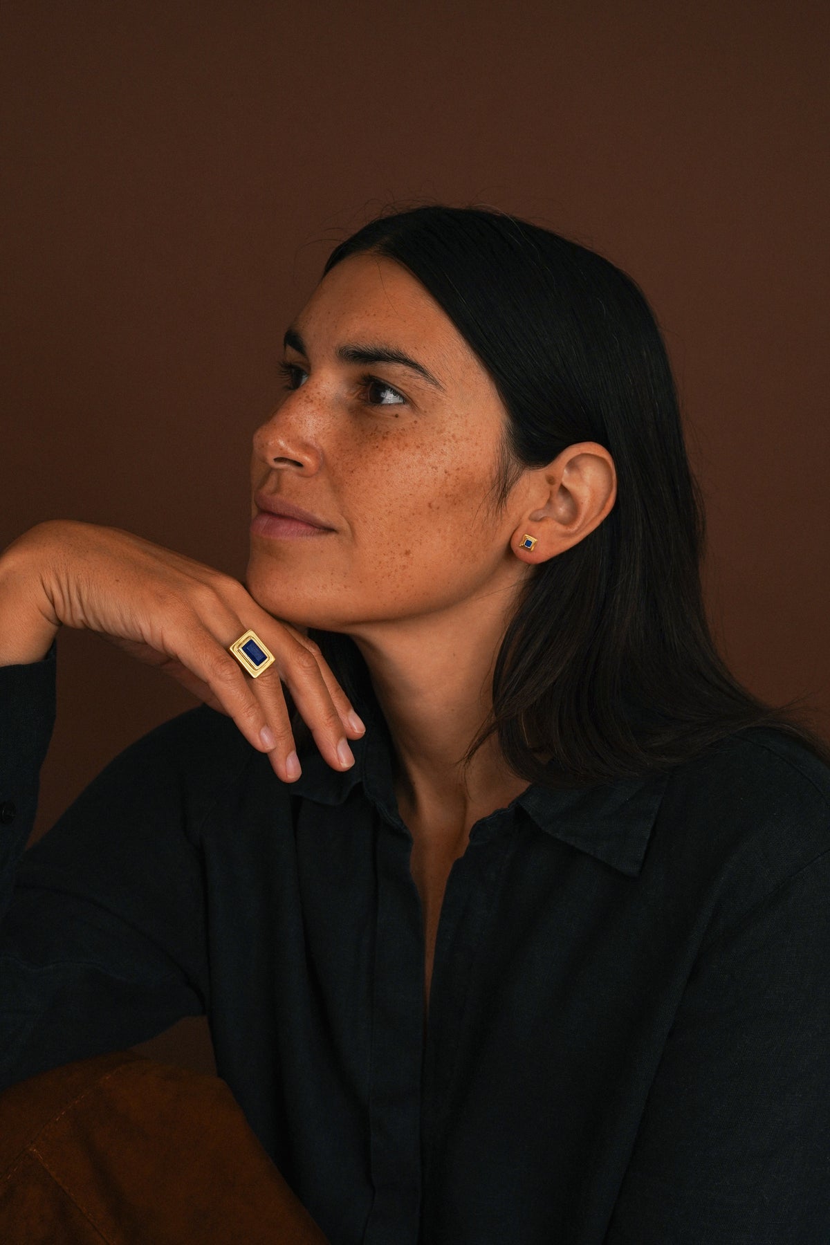 A woman with dark hair wearing a black shirt and a gold ring with a blue stone, posing against a solid dark brown background.