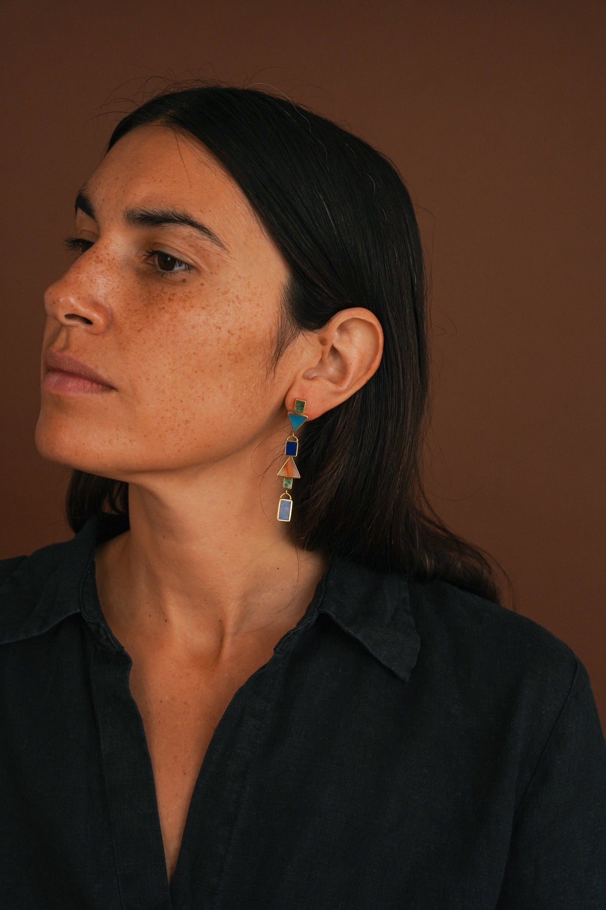 A woman with dark hair wearing a dark navy shirt and a colorful geometric dangle earring, shown in a close-up portrait against a solid brown background.