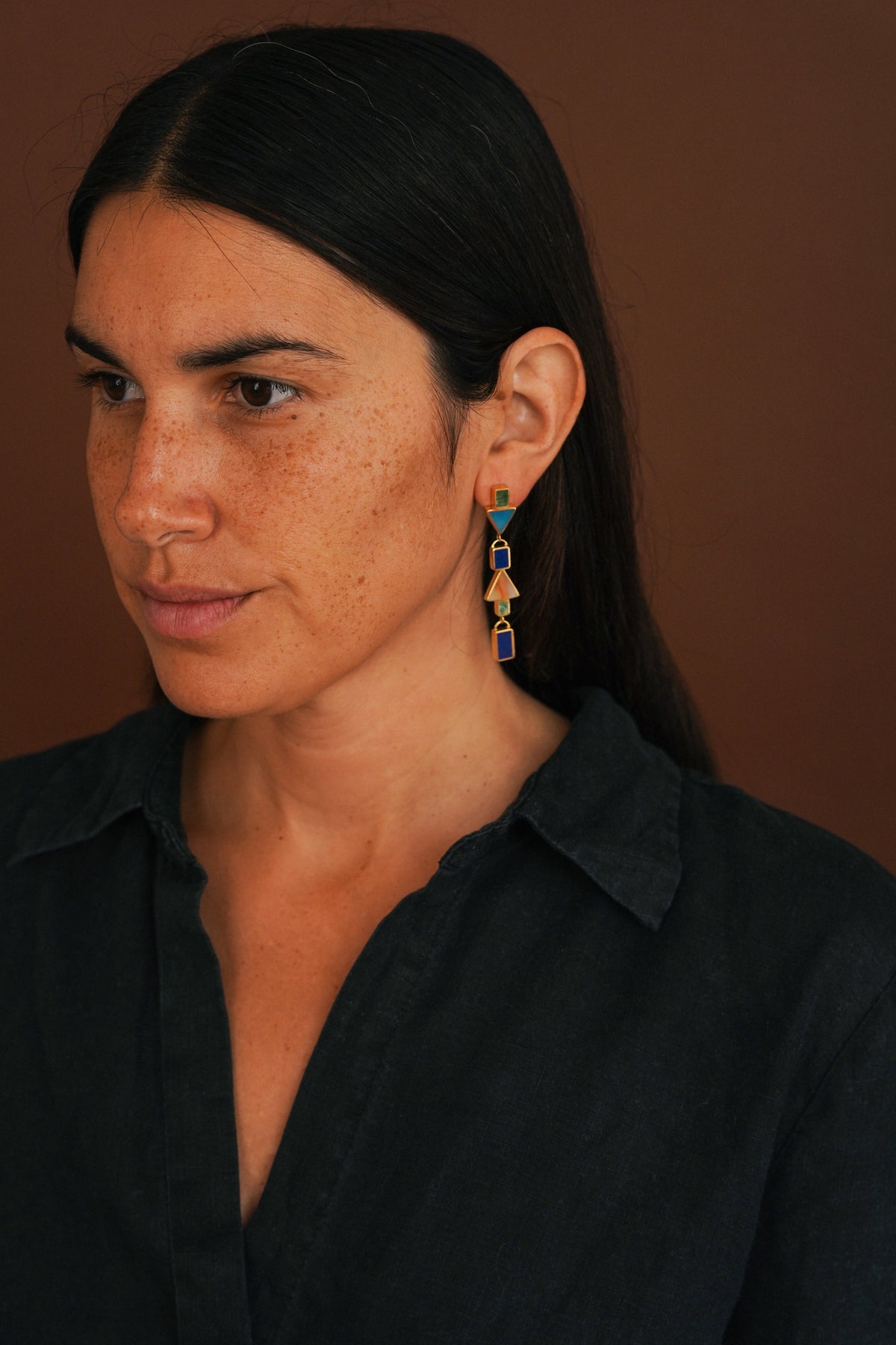 A close-up portrait of a woman with dark hair wearing a black shirt and a colorful beaded drop earring against a warm brown background.