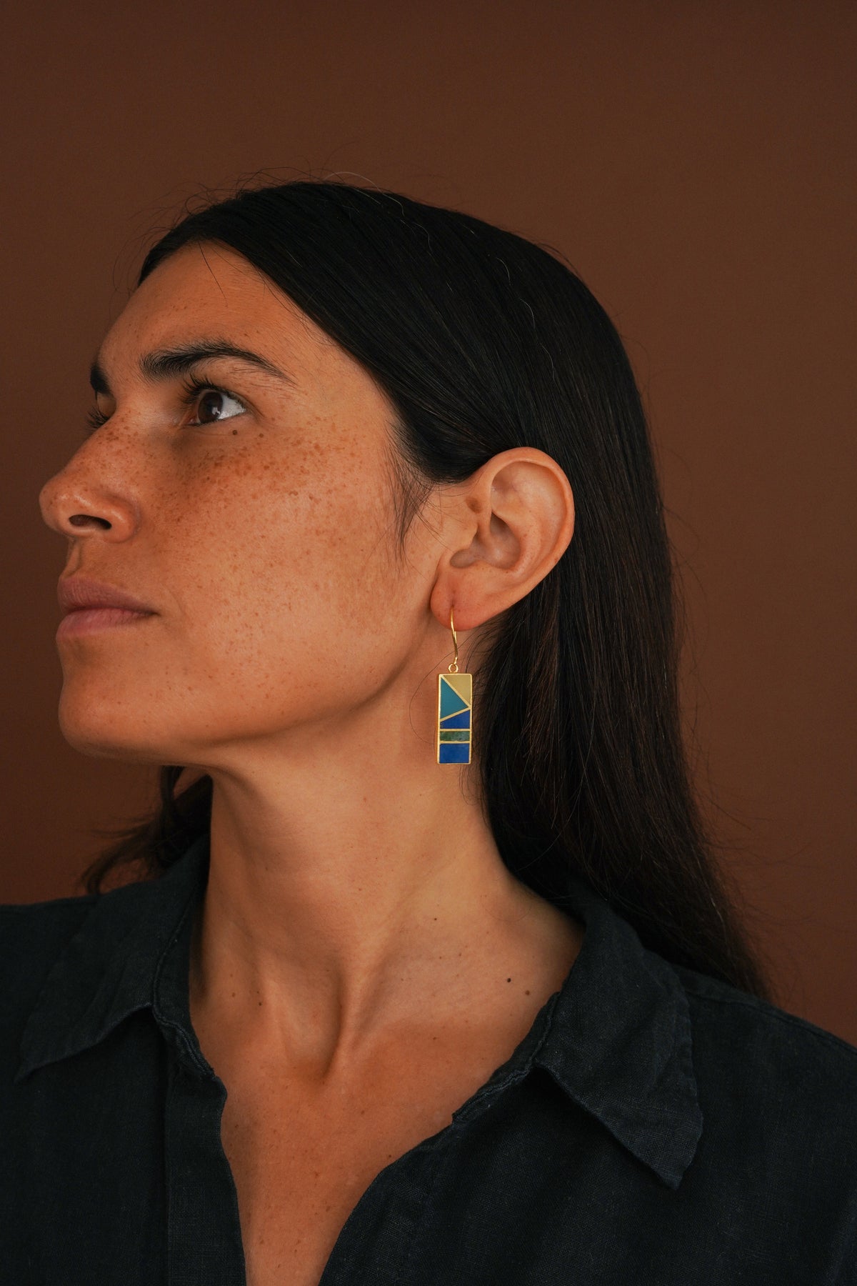 A woman in profile wearing a gold dangle earring with a blue and yellow geometric patterned pendant, set against a solid warm brown background.