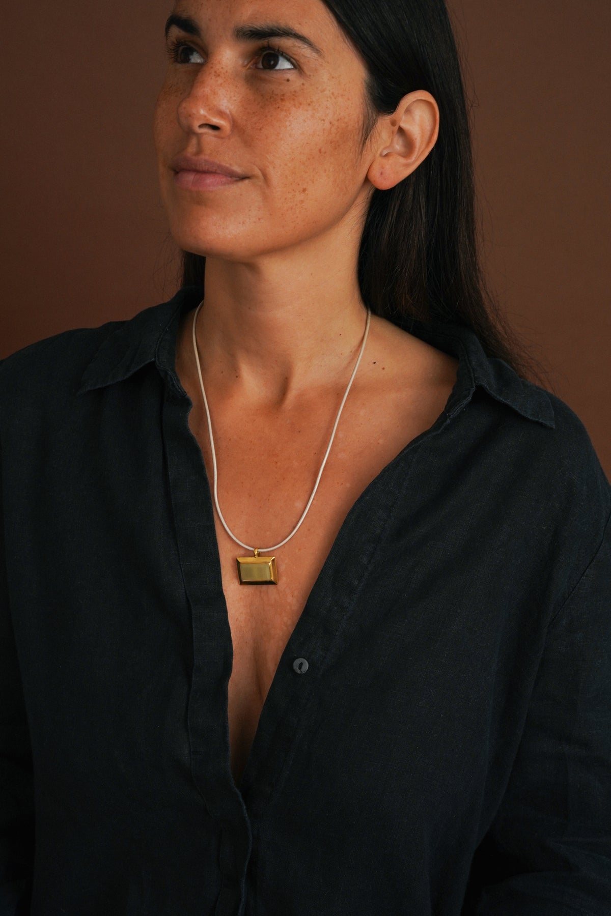 A woman wearing a delicate silver chain necklace with a small gold rectangular pendant, styled with a dark navy linen button-up shirt against a warm brown background.