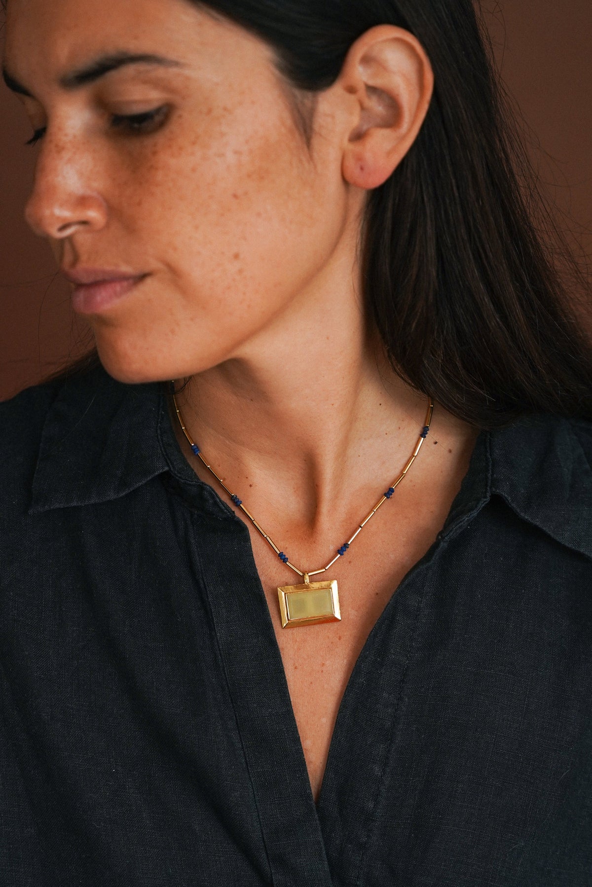 A close-up lifestyle shot of a woman wearing a gold rectangular pendant necklace with dark blue beads on a cord, paired with a dark charcoal shirt against a warm brown background.