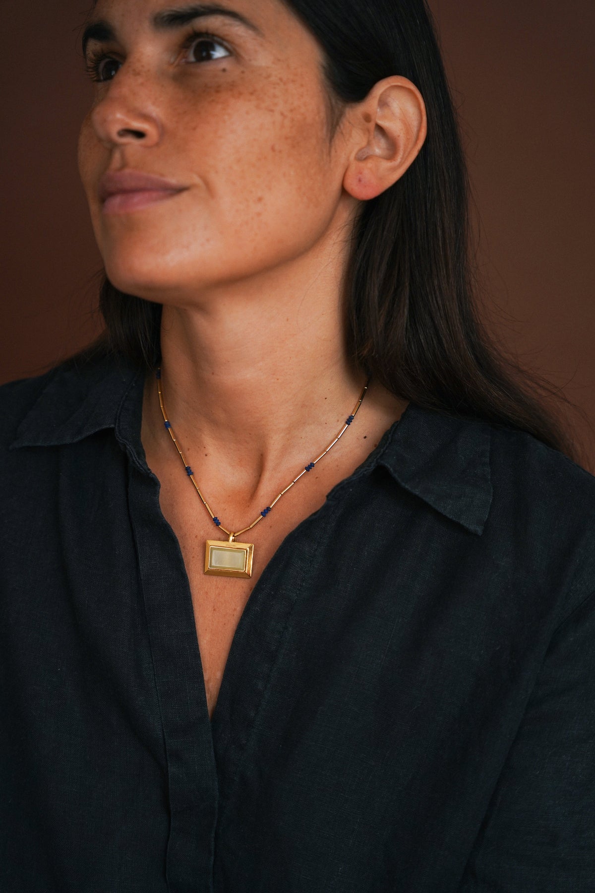 A close-up lifestyle shot of a woman wearing a gold rectangular pendant necklace with blue beads on a dark brown background.
