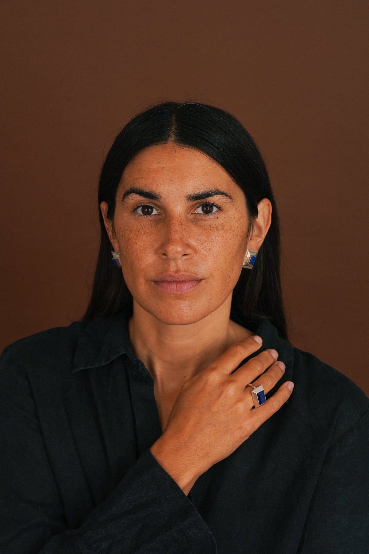 A woman with dark hair and freckles wearing a black shirt, posing with her hand near her neck against a solid brown background. She is wearing a silver ring with a blue stone on her ring finger.