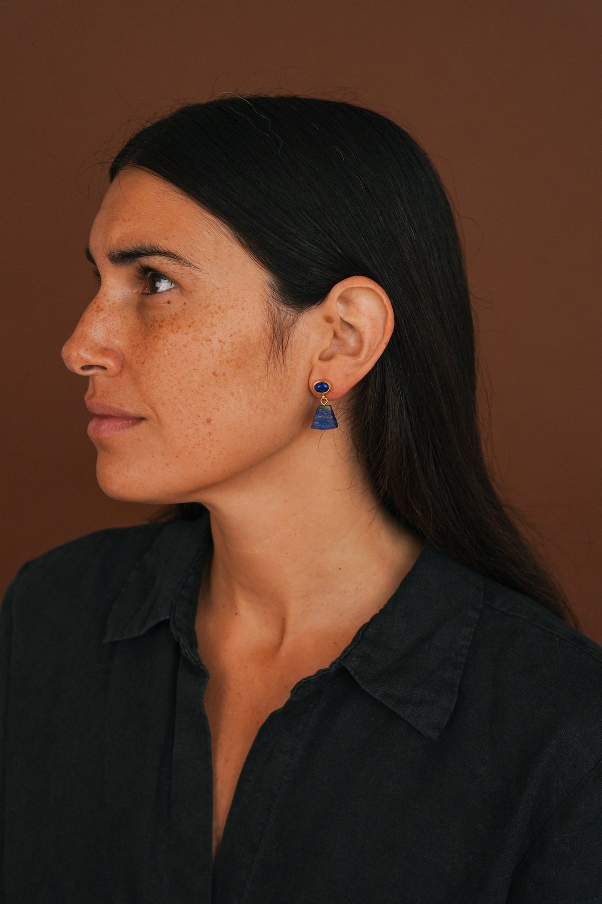A woman in profile wearing a dark navy collared shirt and a small blue geometric drop earring, set against a solid warm brown background.