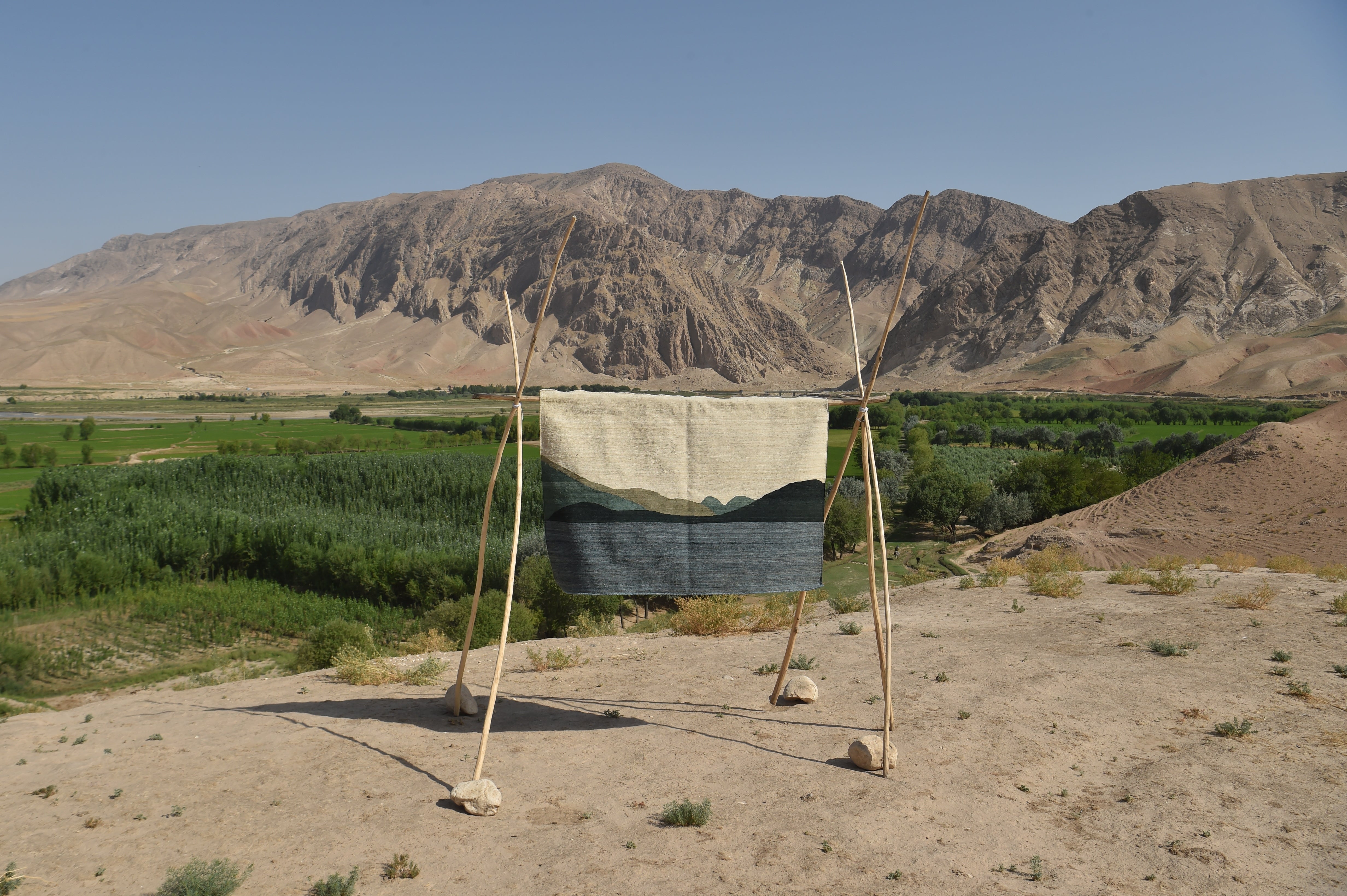A woven textile with a mountain landscape design hangs on a simple wooden frame outdoors, with green fields and large tan mountains in the background under a clear blue sky.