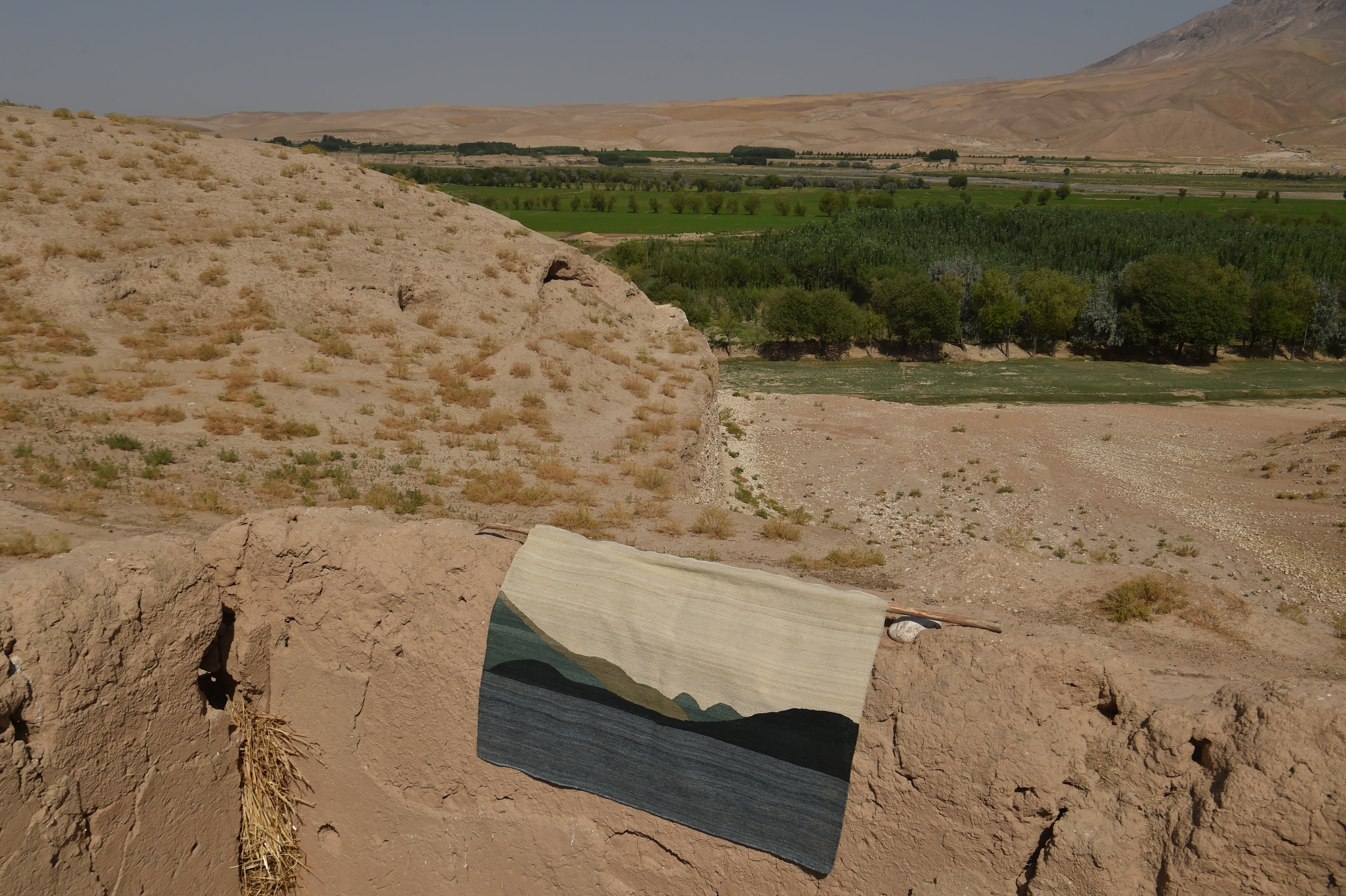 A rectangular flatweave rug with a landscape design of mountains and water, draped over a rustic earthen wall in a dry, rural outdoor setting with hills and a green field in the background.
