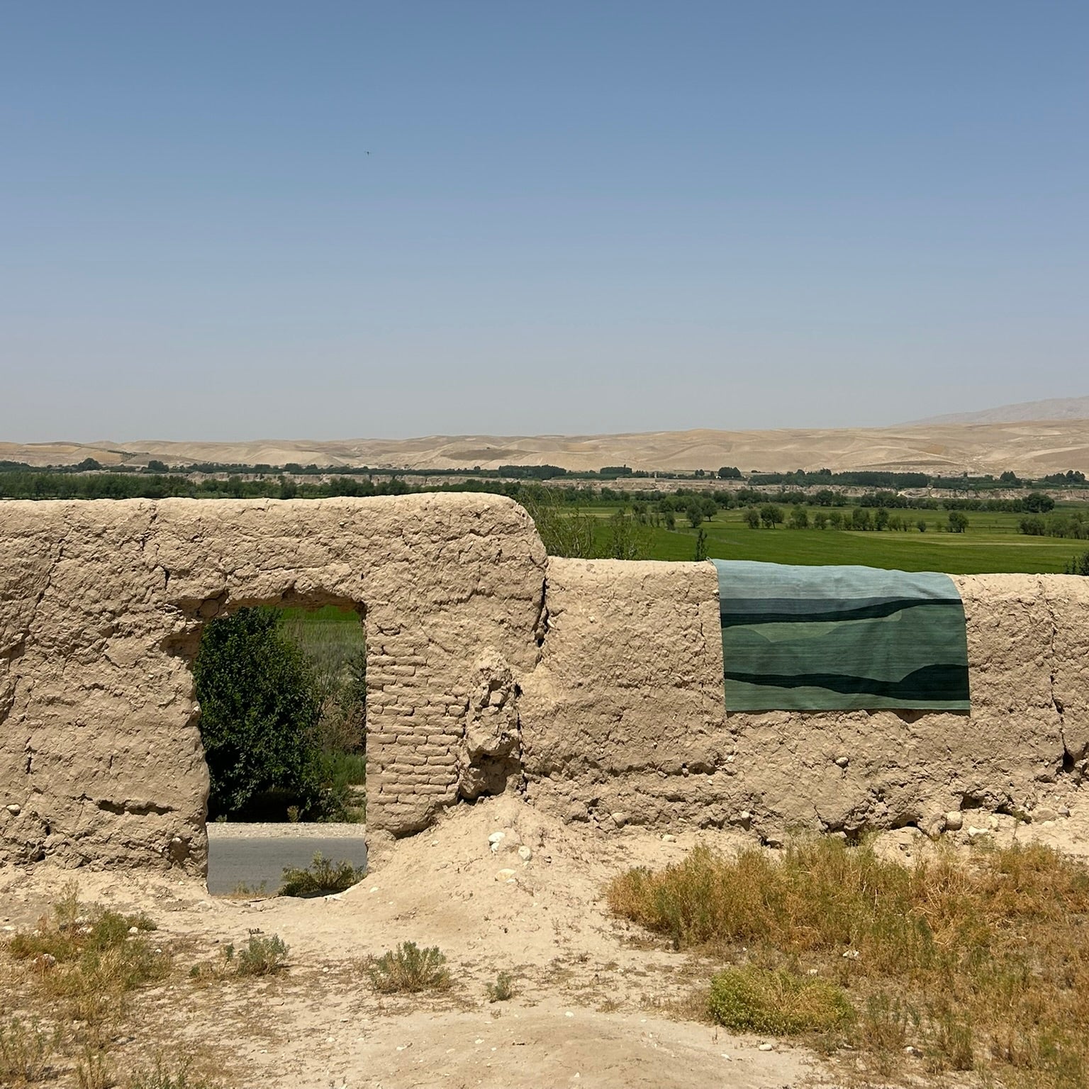 A wide outdoor landscape with a sandy adobe wall in the foreground, a green tarp or fabric panel on the right, and a distant green valley under a clear blue sky.