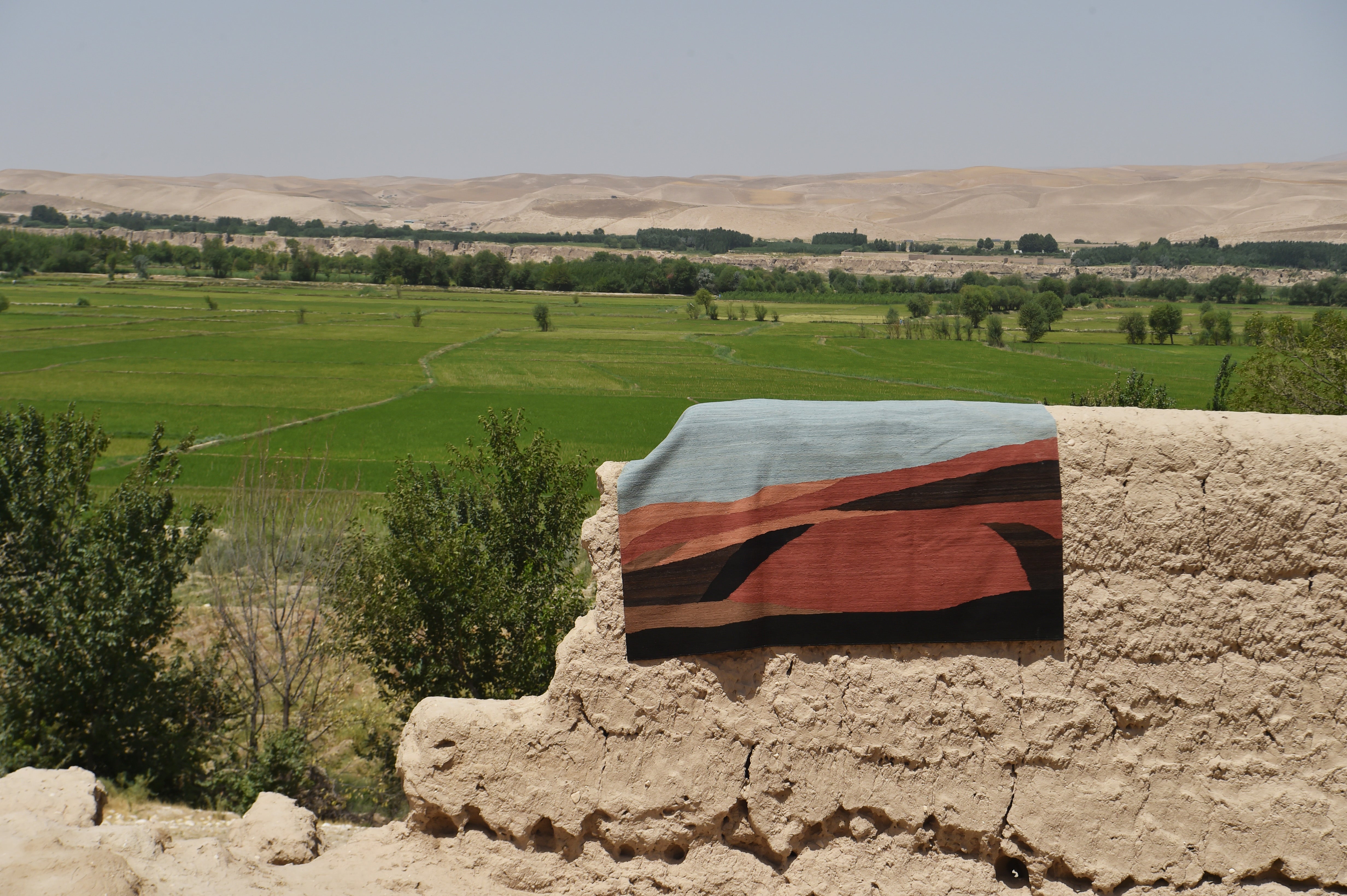 A woven rug with a geometric pattern in light blue, terracotta red, black, and brown is draped over a rustic beige mudbrick wall, with a lush green valley and desert hills in the background.
