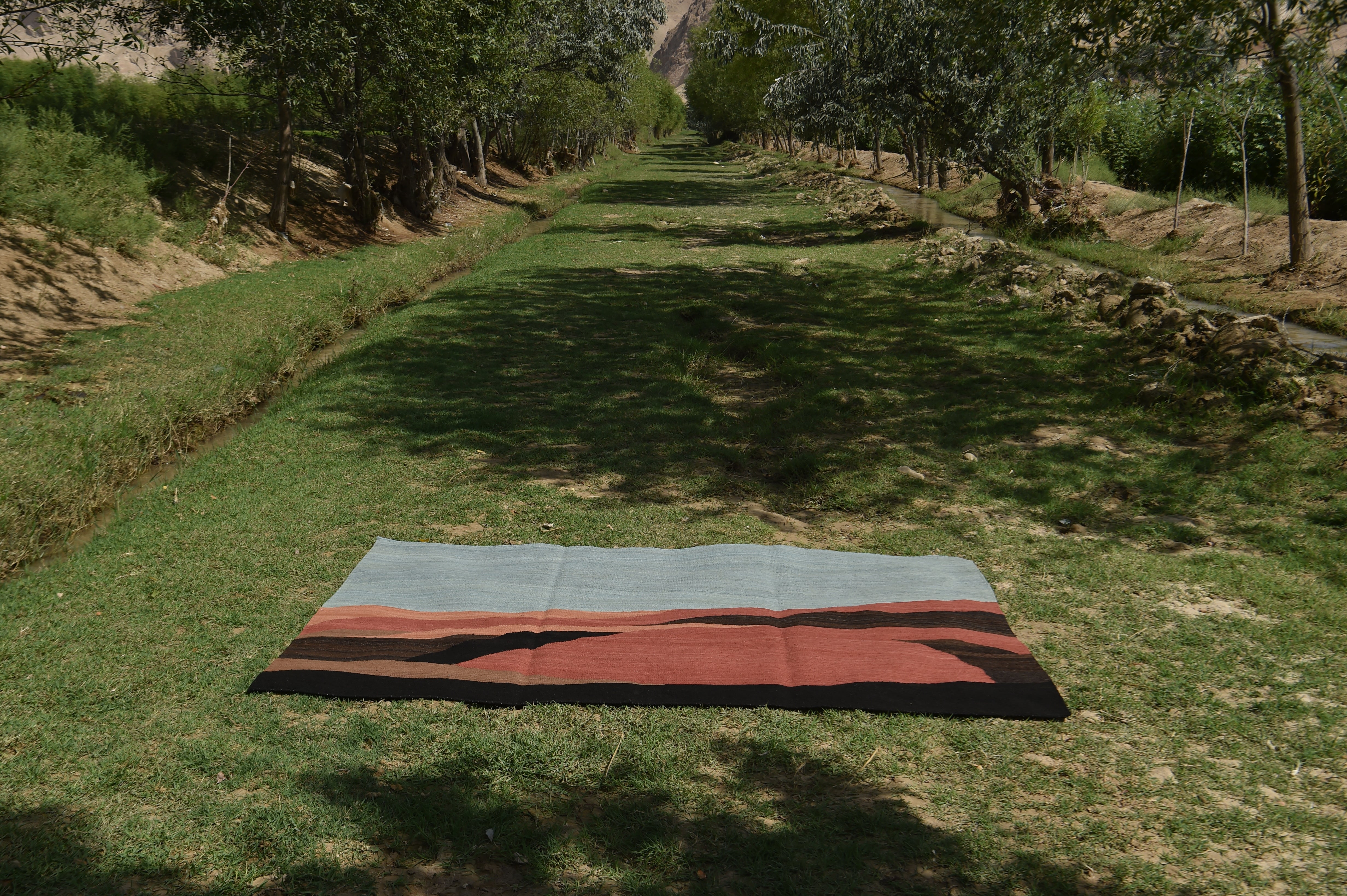 A rectangular flatweave rug with a geometric mountain pattern in light blue, rust red, and black, laid out on a grassy path between rows of trees.