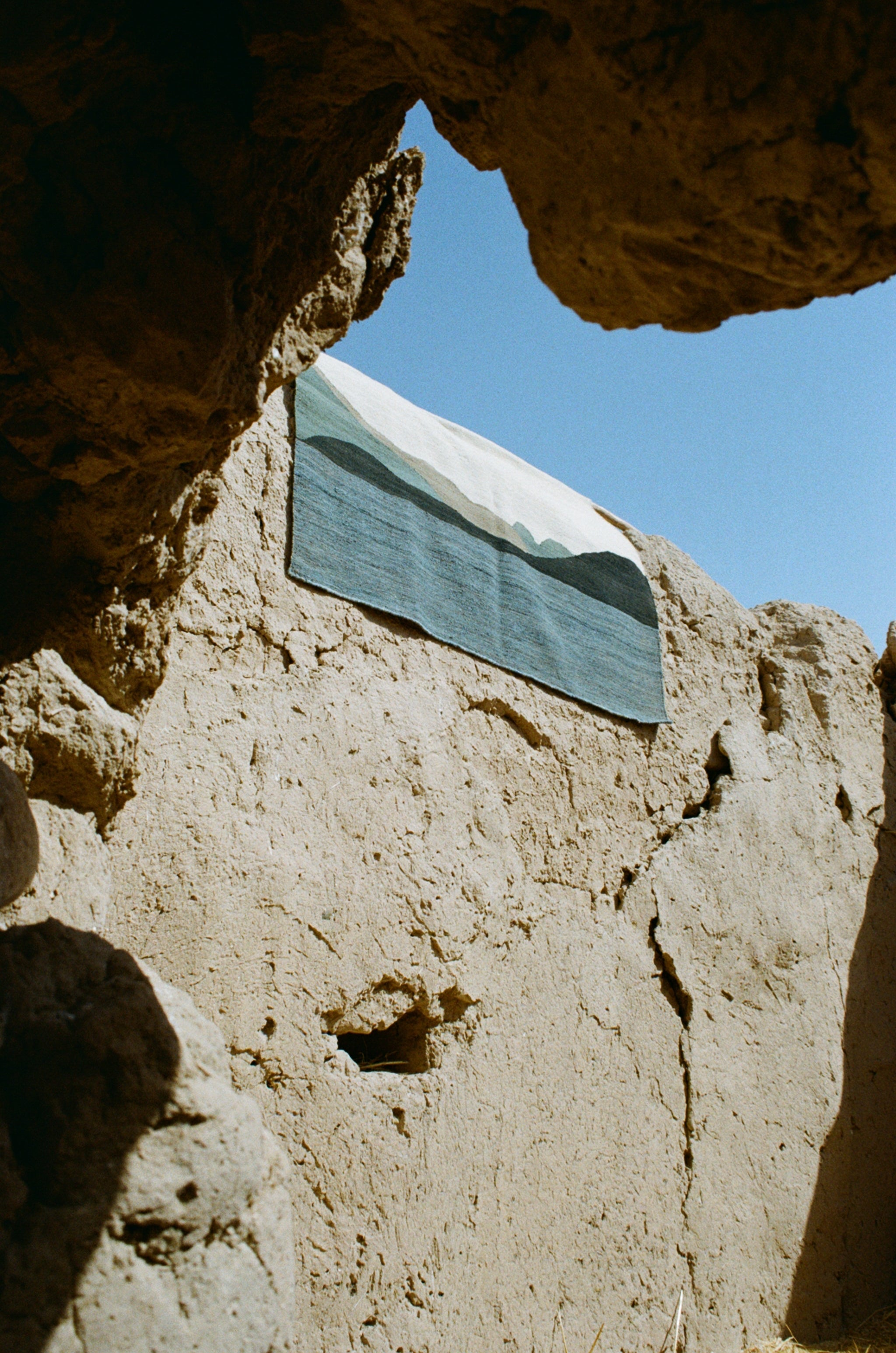 A rectangular textile rug with a blue and white mountain landscape design, draped against a rustic adobe wall under a clear blue sky.