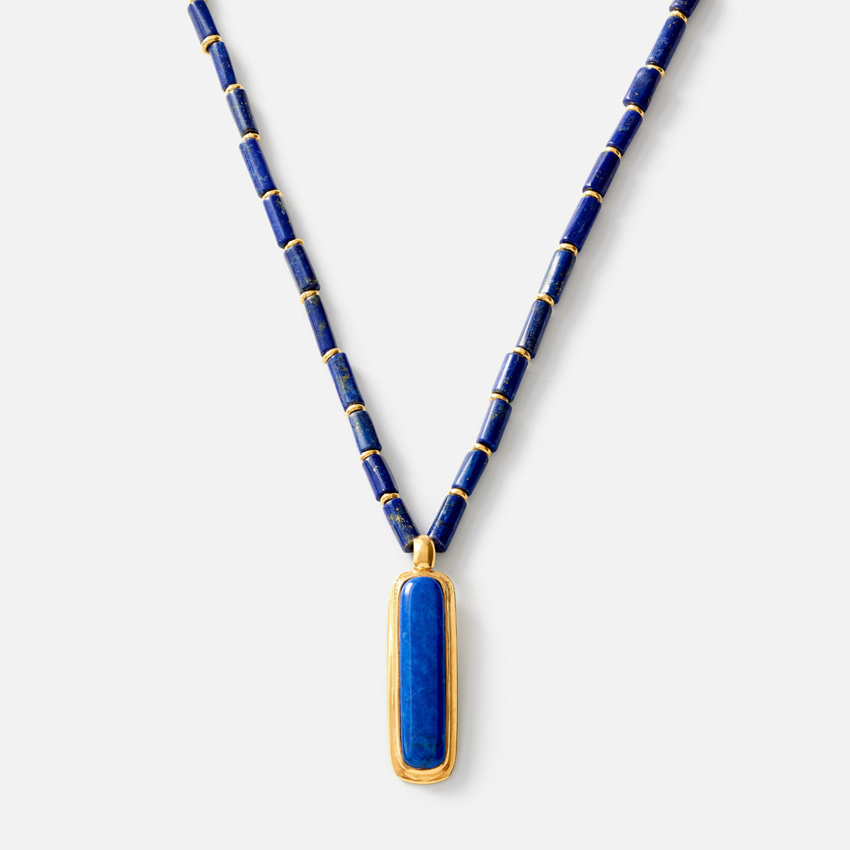 A luxury necklace featuring a deep blue lapis lazuli pendant in a gold bezel setting, suspended from a matching blue beaded chain with gold spacer rings, shot against a light gray background.