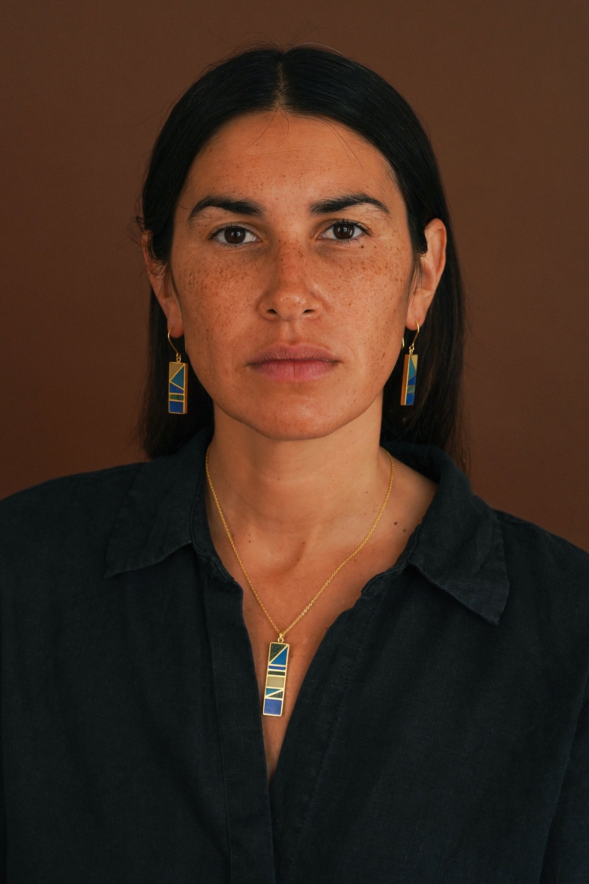 A woman with dark hair pulled back wears a gold necklace and matching drop earrings with blue and yellow geometric inlay details, set against a solid brown background.