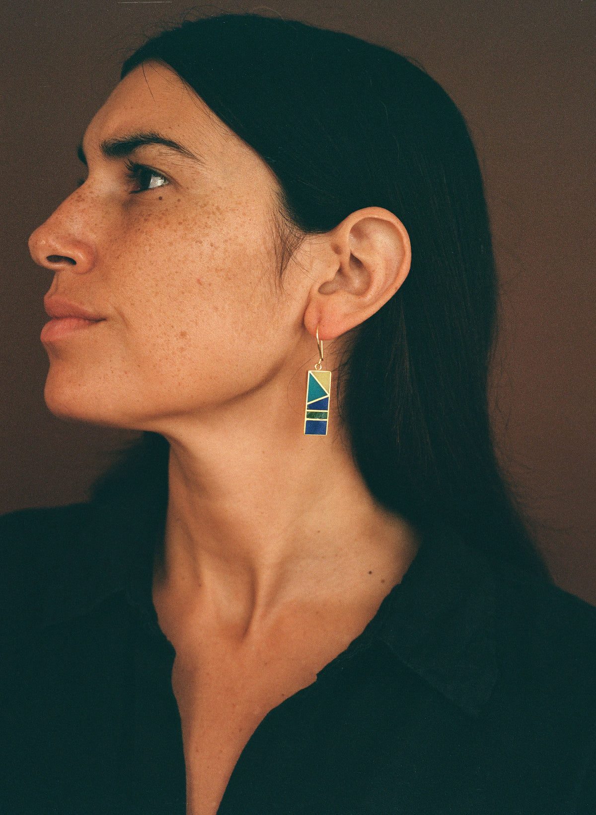 A woman in profile wearing a geometric drop earring with blue, yellow, and gold triangular patterns, set against a dark brown background.