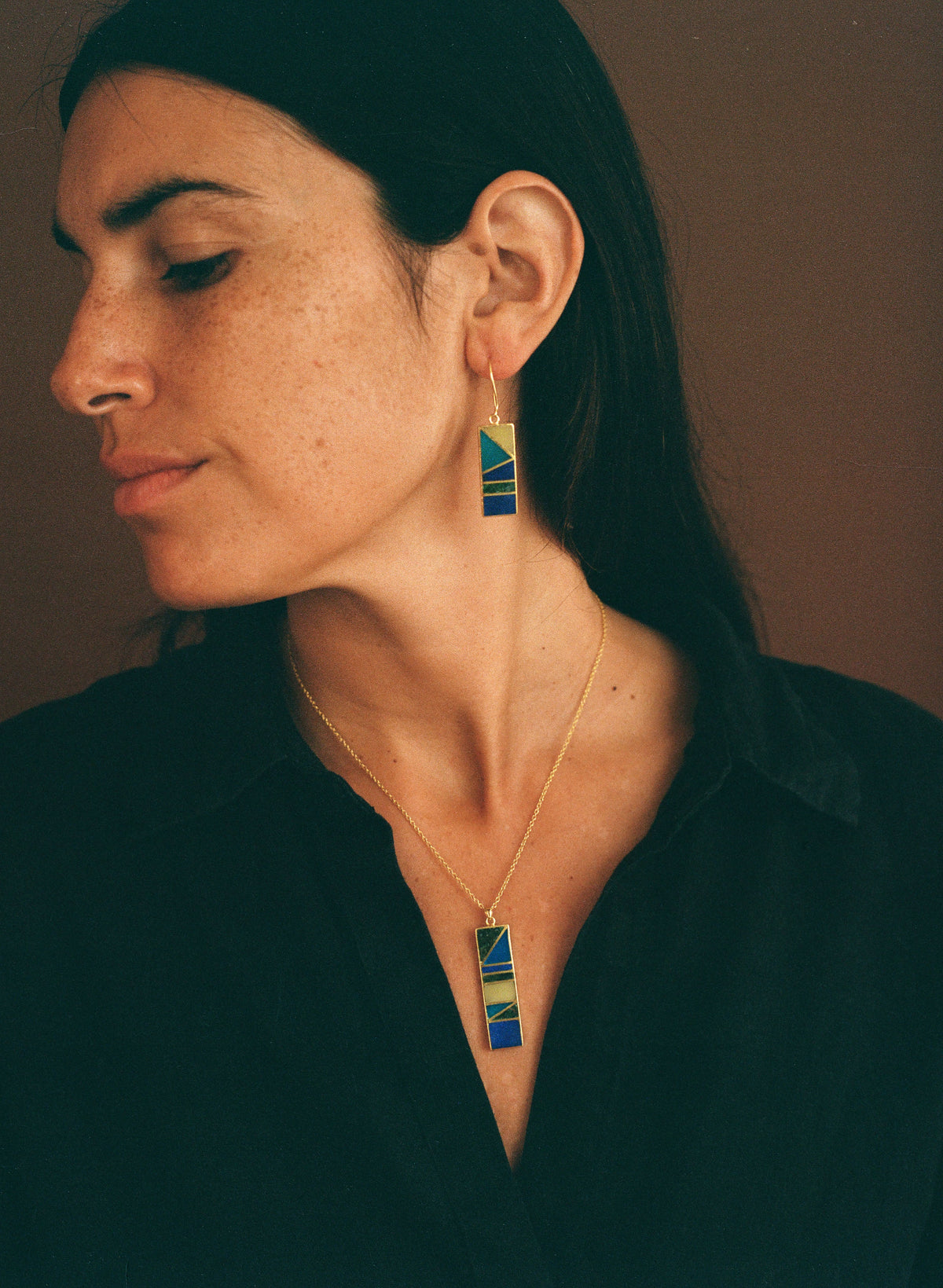 A woman wearing a gold necklace and matching drop earring with rectangular blue, green, and yellow geometric enamel inlays, shown in a close-up portrait against a dark brown background.