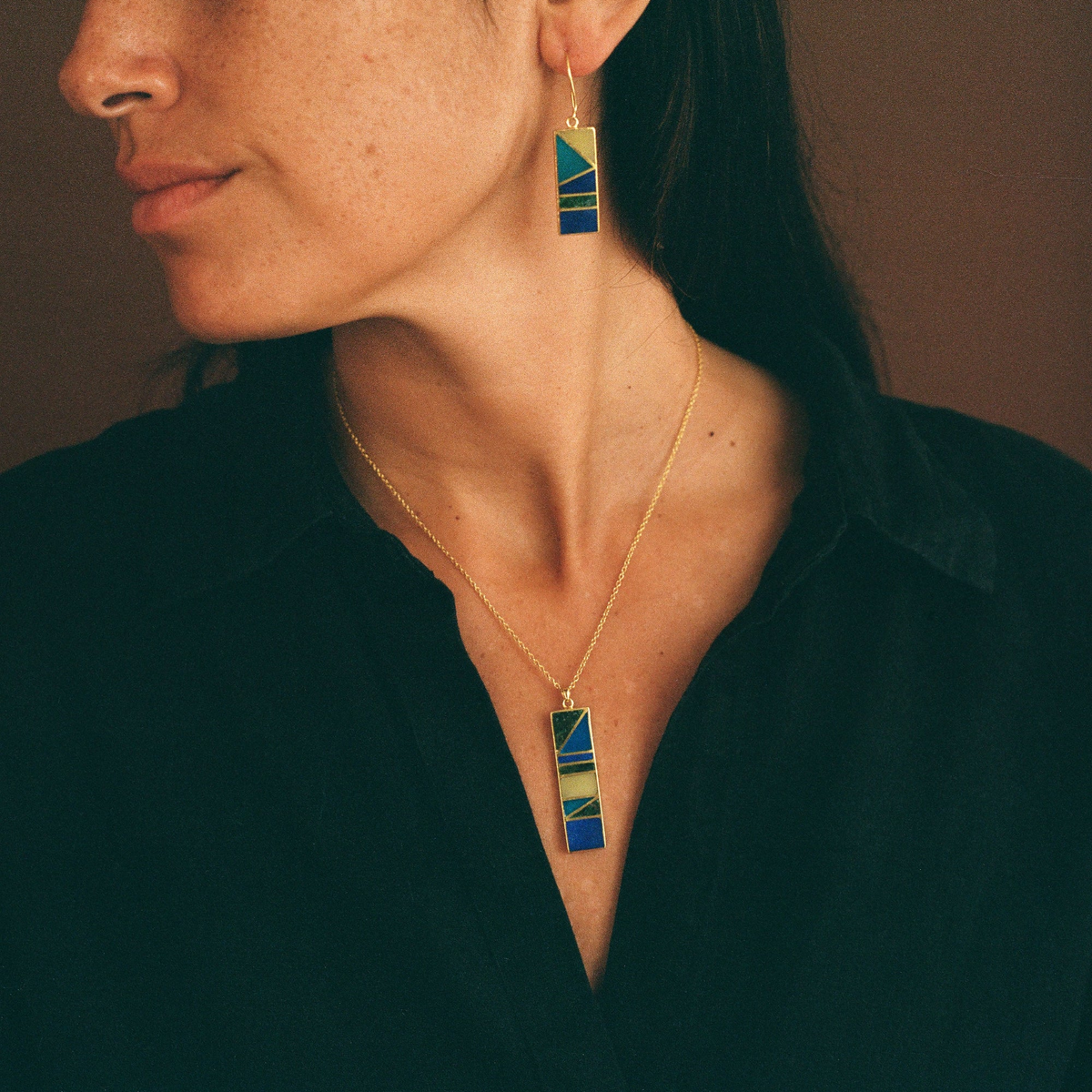 Close up of woman with pendant necklace and dangling earrings of lapis lazuli stones. wearing a dark shirt and a soft brown background.