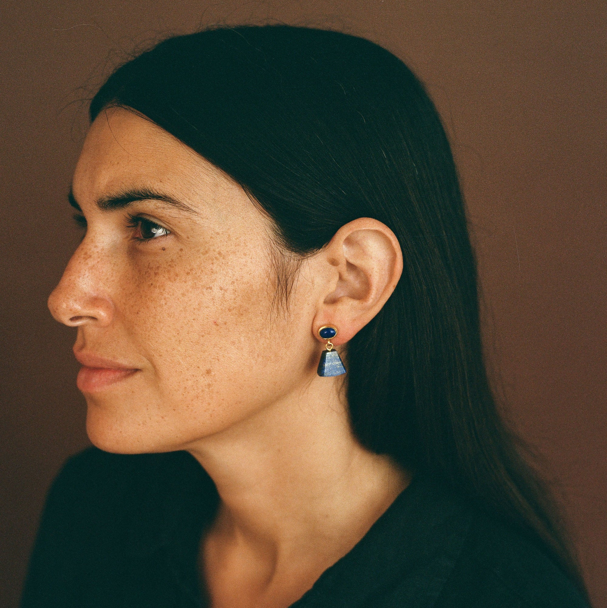 A close-up side profile of a woman with dark hair wearing a small geometric dangle earring with a dark blue top and a light blue triangular drop, against a warm brown background.