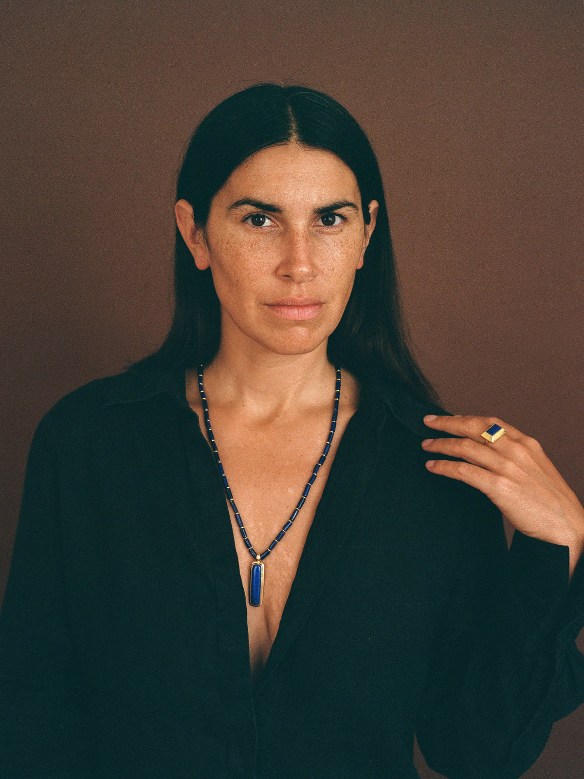 A woman with long dark hair wearing a black shirt and a beaded necklace with a blue pendant, posing against a solid brown background.