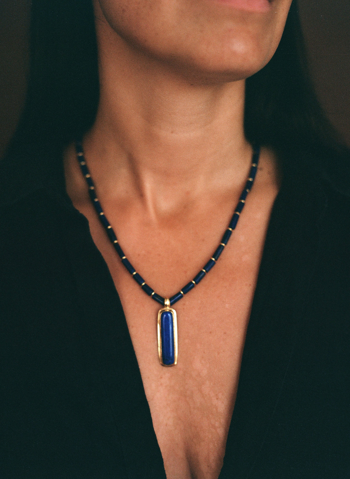A close-up of a woman wearing a beaded necklace with a rectangular blue pendant in a gold frame.