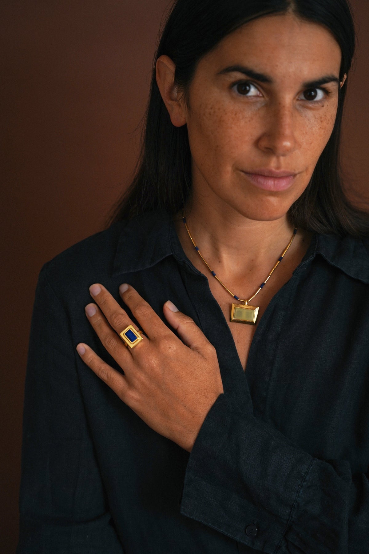 A woman wearing a gold ring with a blue square stone on her finger, paired with a gold necklace, against a dark brown background.