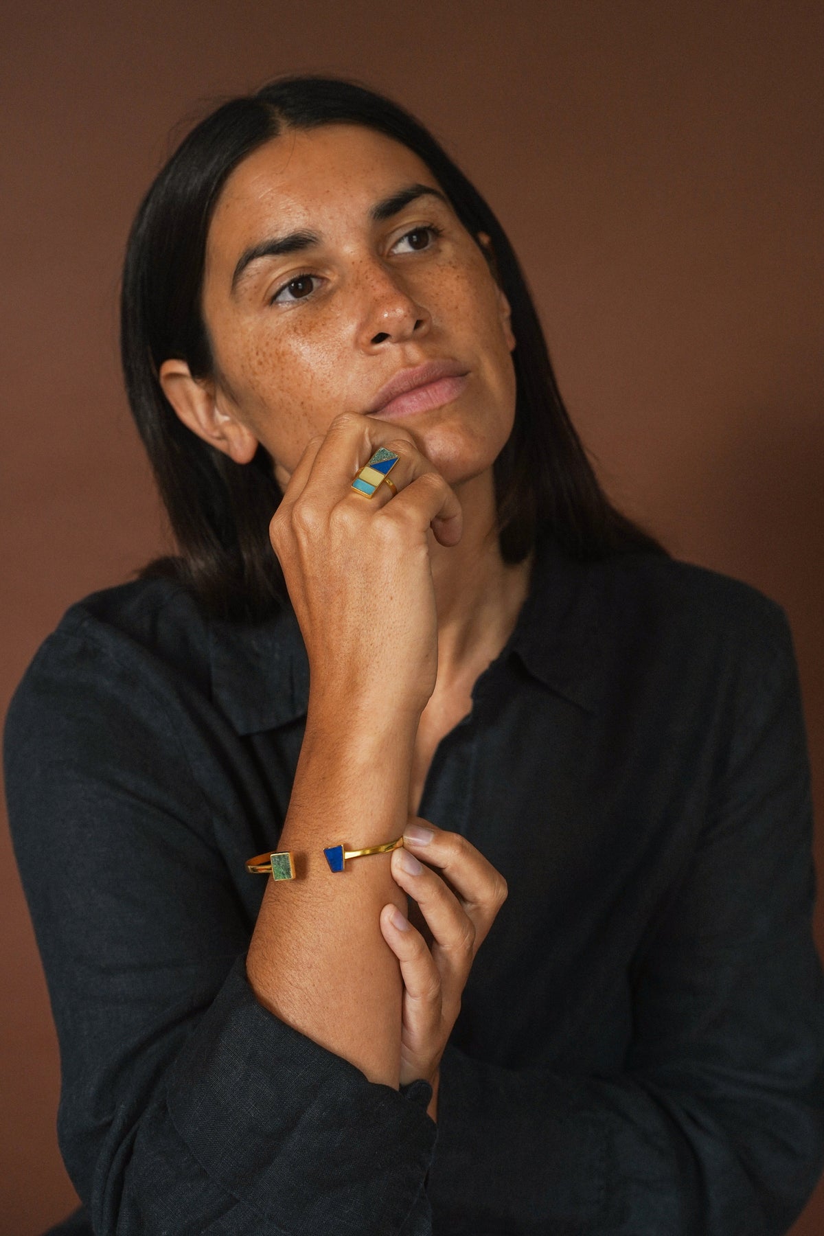 A woman with dark hair wearing a black shirt, resting her chin on her hand. She is wearing a gold-toned bracelet with blue and green rectangular accents on her wrist and a matching ring on her finger, set against a solid brown background.
