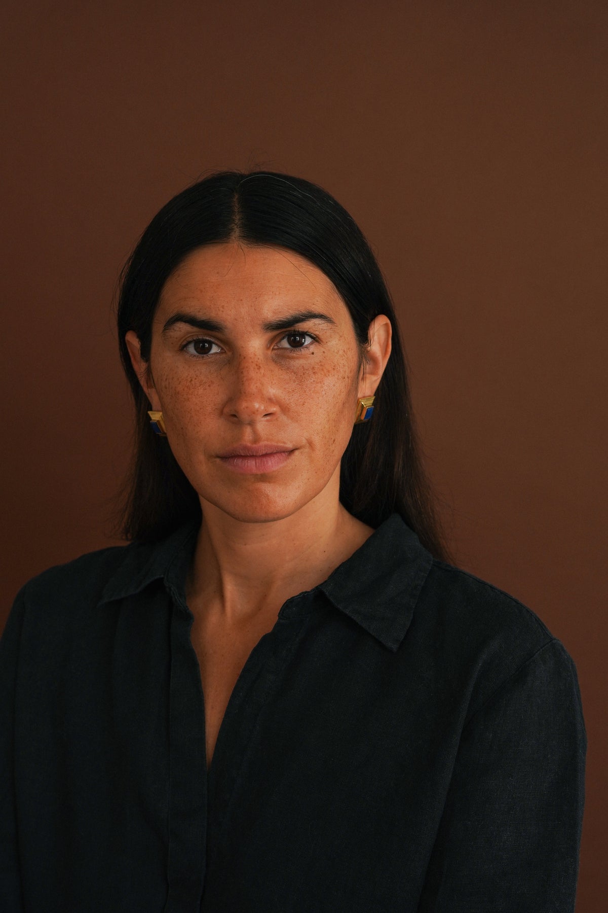 A woman with dark hair and freckles wearing a dark navy button-up shirt and small gold hoop earrings, photographed against a solid warm brown background.