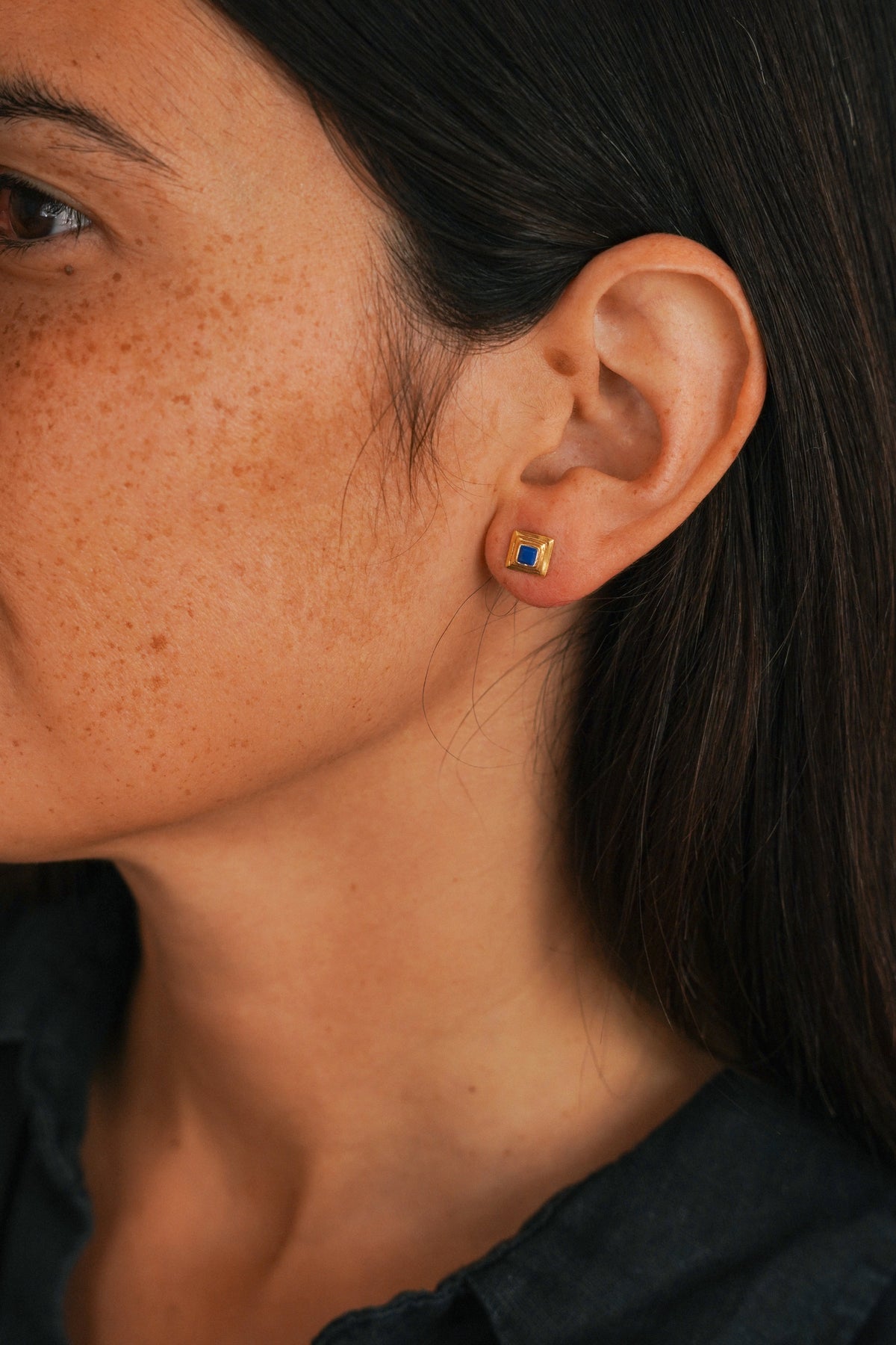 Close-up of a woman's ear wearing a small square gold earring with a blue and red inset, against dark hair and skin.