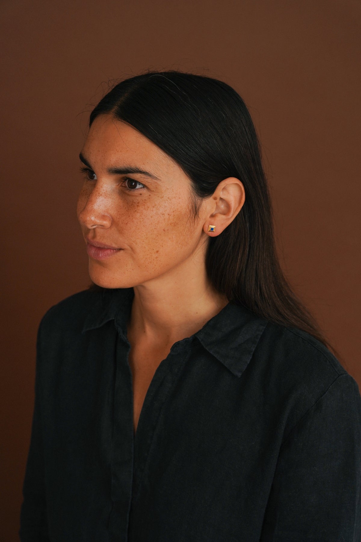 A woman with long dark hair wearing a dark button-up shirt and a small gold stud earring, shown in a side profile against a solid brown background.