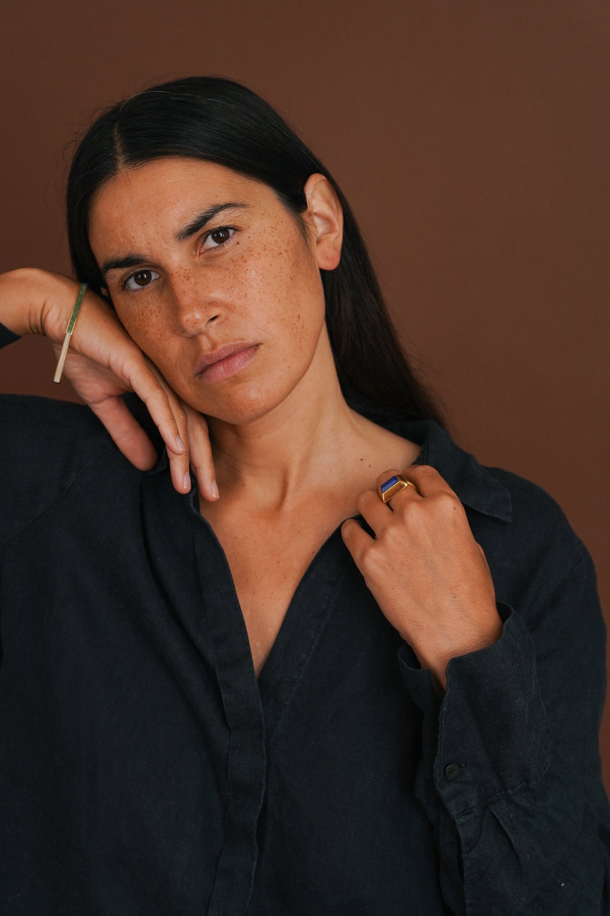 woman with dark hair models a gold bangle and ring, wearing a dark shirt and a brown background