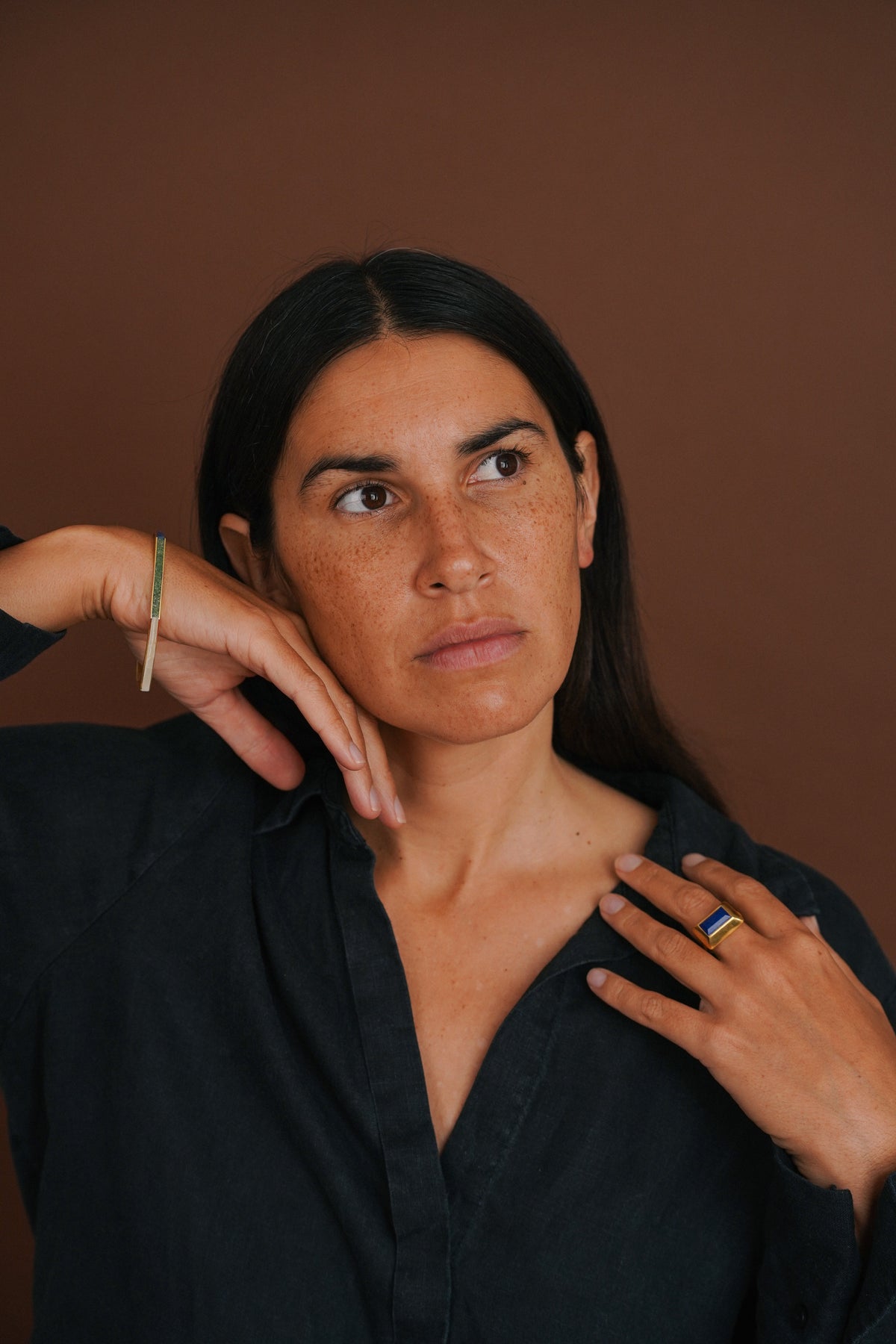 A woman with dark hair wearing a black linen shirt, posing with her hands near her face against a solid brown background.