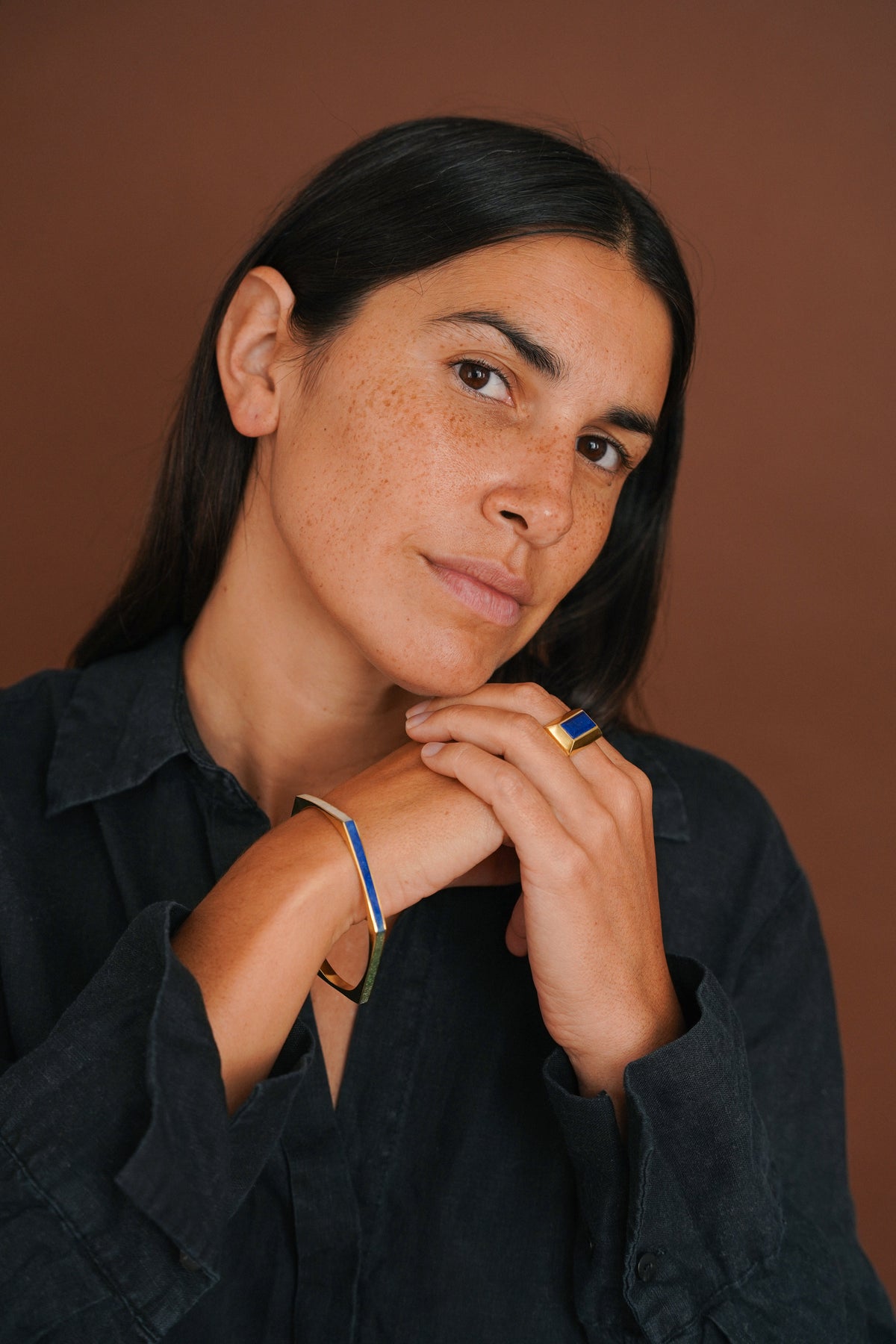A close-up portrait of a woman with dark hair and freckles, wearing a black shirt and jewelry including a gold ring with a blue stone and a thin gold bracelet, set against a solid brown background.