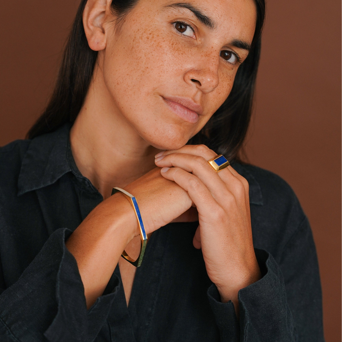 A close-up lifestyle shot of a woman wearing a gold-plated bangle bracelet with blue enamel stripes and a matching gold ring with a blue rectangular stone, set against a solid warm brown background.
