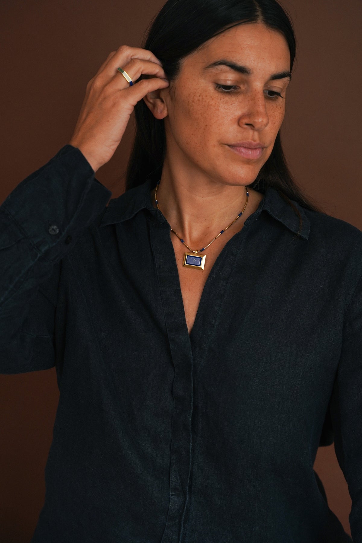 A woman wearing a dark navy blue button-up shirt and a silver-toned necklace with a rectangular blue and white pendant, posing against a solid dark brown background.