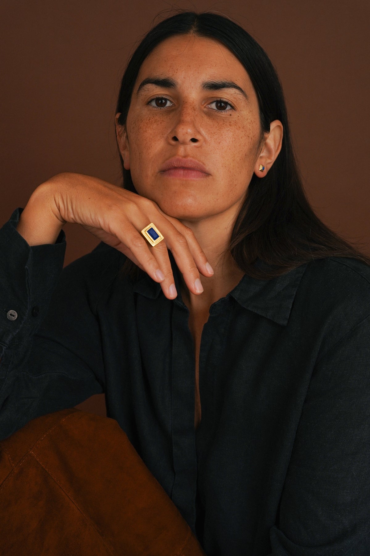 A woman with dark hair resting her chin on her hand, wearing a black button-up shirt and a gold ring with a blue rectangular stone, set against a warm brown background.