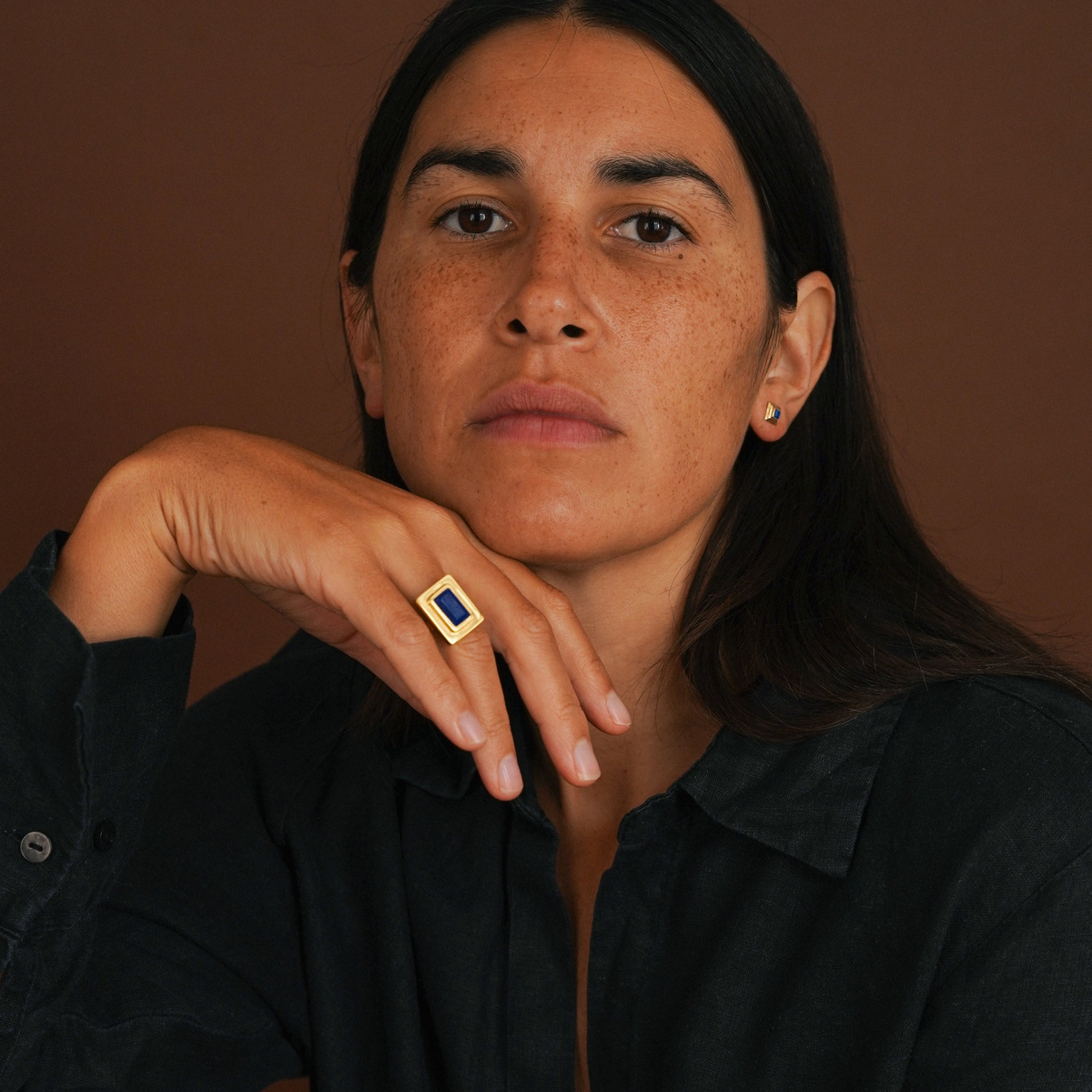 A close-up portrait of a woman with dark hair resting her chin on her hand, wearing a gold ring with a square blue stone. She is wearing a dark button-down shirt against a solid brown background.