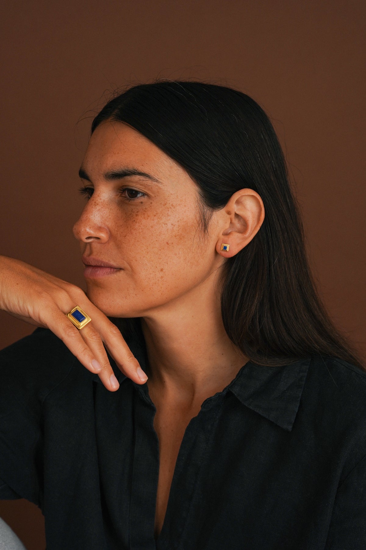 A woman with dark hair wearing a black shirt, shown in profile with her hand resting near her face. She is wearing a gold ring with a blue rectangular stone on her ring finger and a small matching blue stud earring.